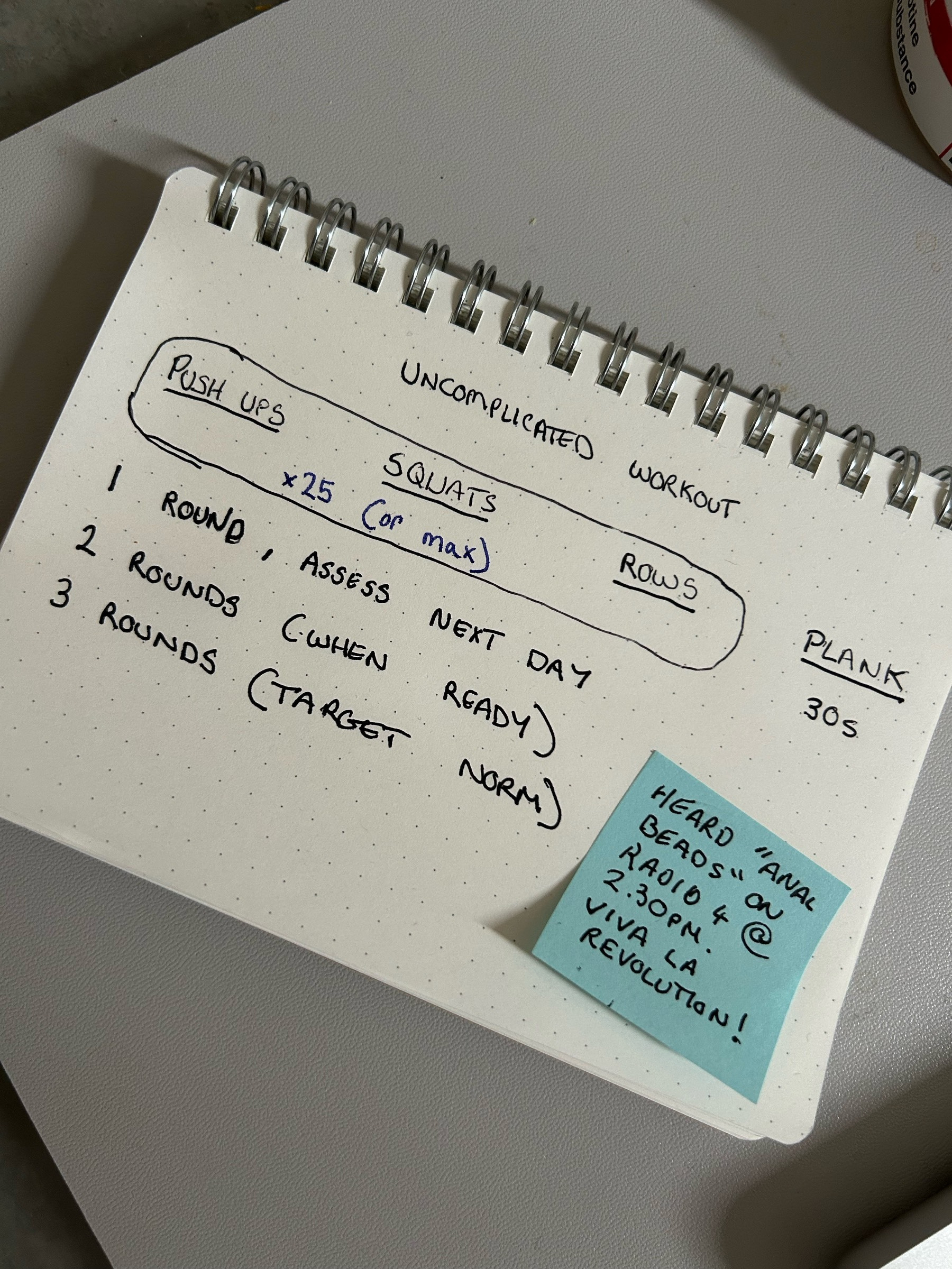 Auto-generated description: A handwritten workout plan titled Uncomplicated Workout outlines exercises including push-ups, squats, rows, and planks, with rounds and assessment instructions, accompanied by a blue sticky note with a reminder about a radio show.