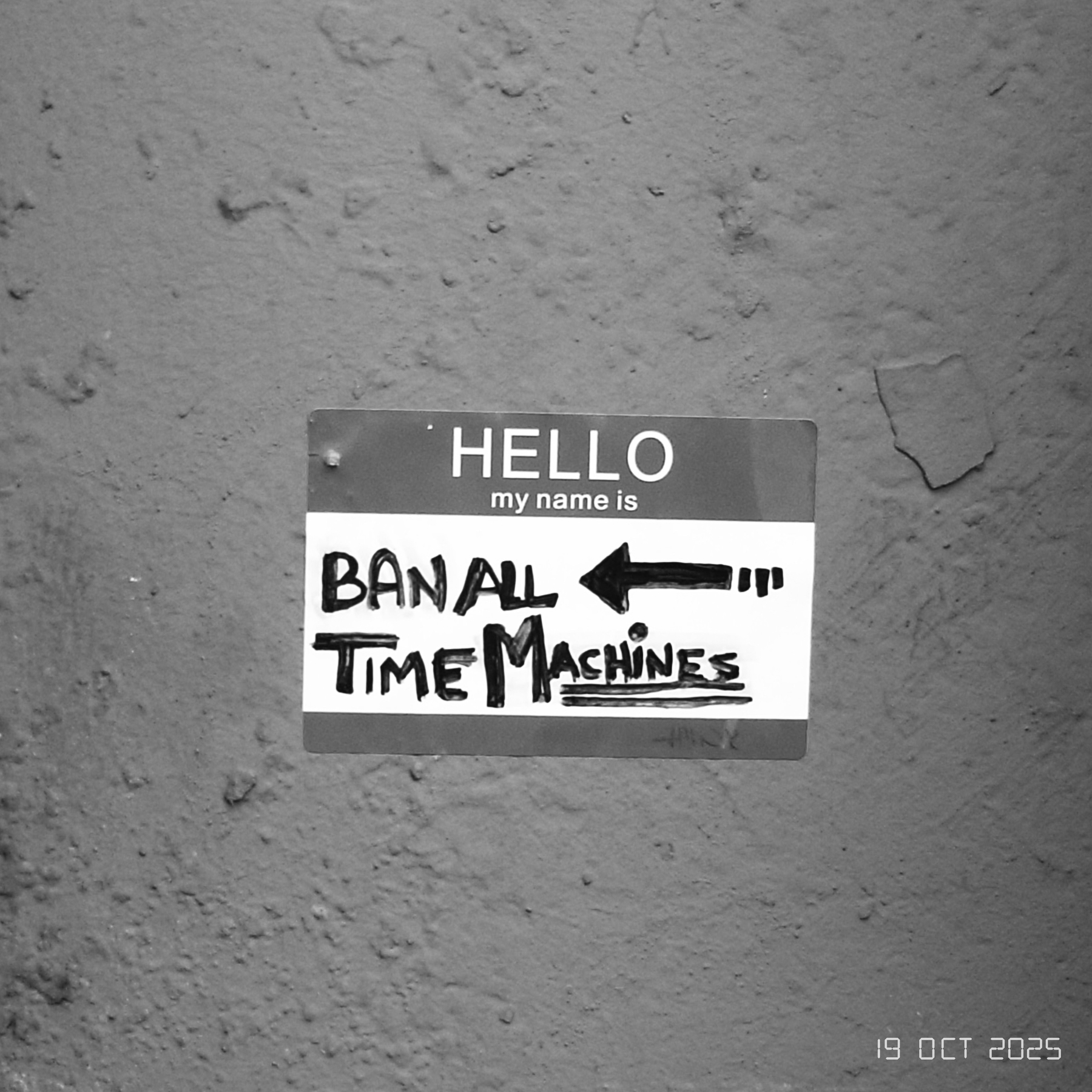 A Hello my name is sticker with Ban All Time Machines written on it is stuck to a textured wall.