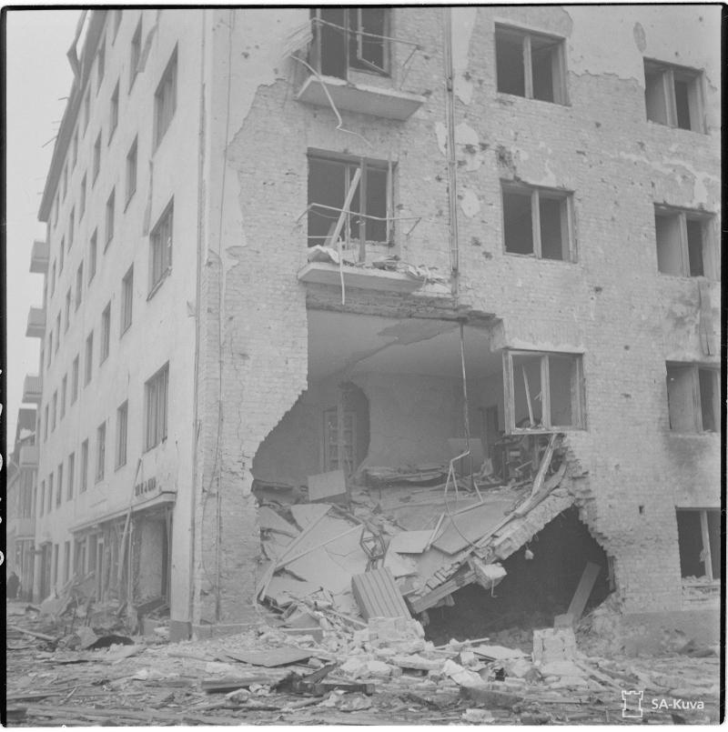 Black and white photo. A corner of a five-floor residential building with a large piece of the wall missing and several windows blown out from an explosion or bomb