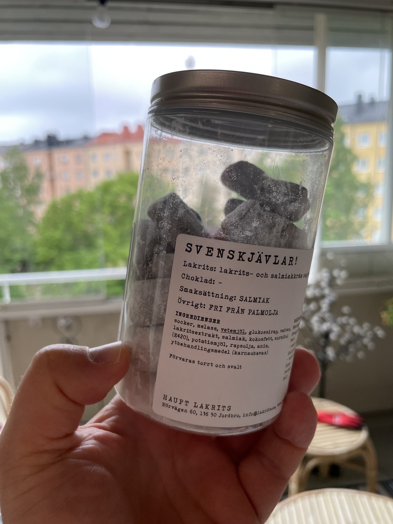 A hand holding a jar of black, powder-coated candies with a white label featuring the large text "SVENSKJÄVLAR!" in Swedish. The background shows an outdoor scene with green trees and several buildings, indicating the jar is being held near a window.