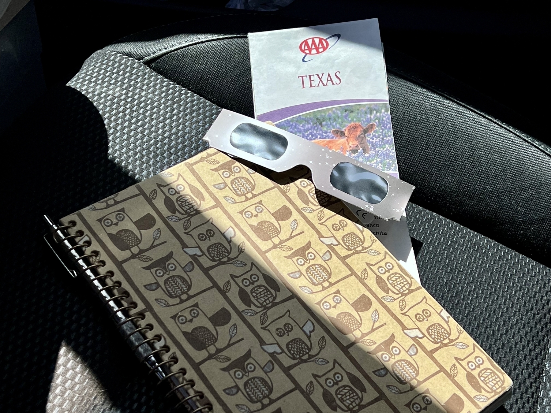 Passenger seat of a car with a notebook, map of Texas, and a pair of disposable solar eclipse safety glasses.