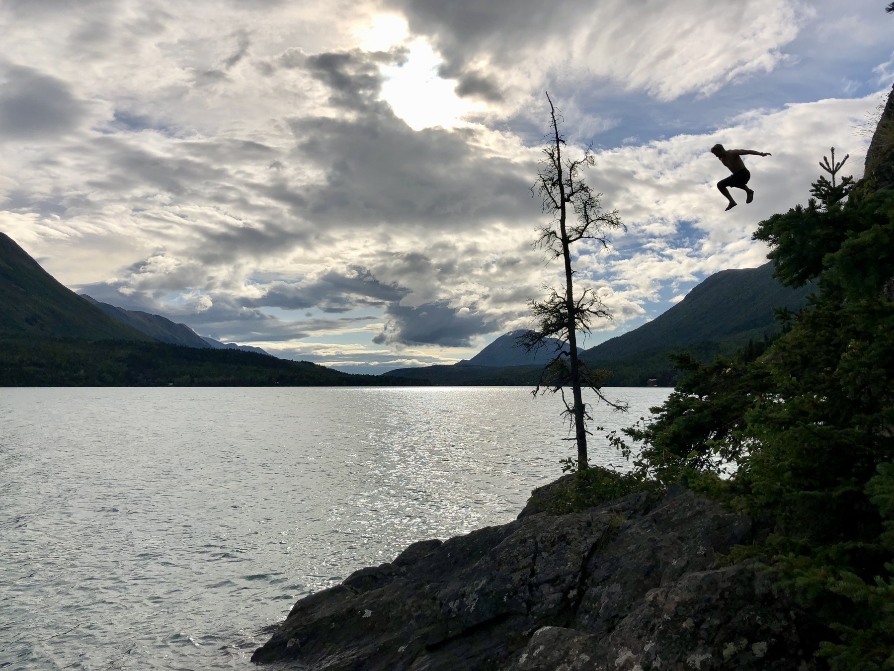 A person is captured mid-jump off a rocky ledge into a body of water, surrounded by mountains and trees under a cloudy sky.