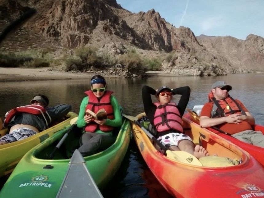 Four individuals in kayaks on the Colorado River; three are napping, one is reading a book.