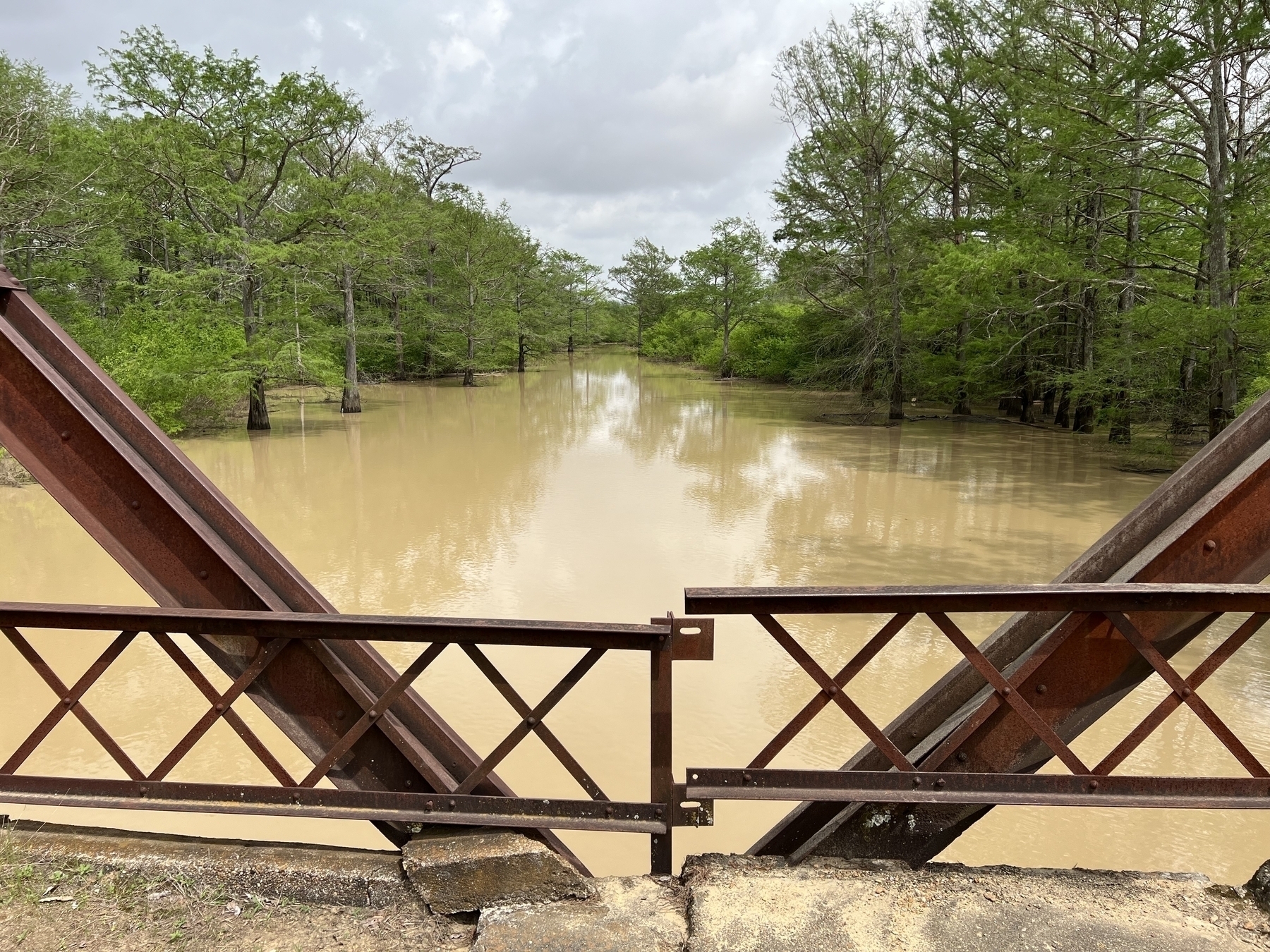 A dilapidated rusted bridge over a flooded bayou, with trees flooded along the banks and muddy water in the channel.