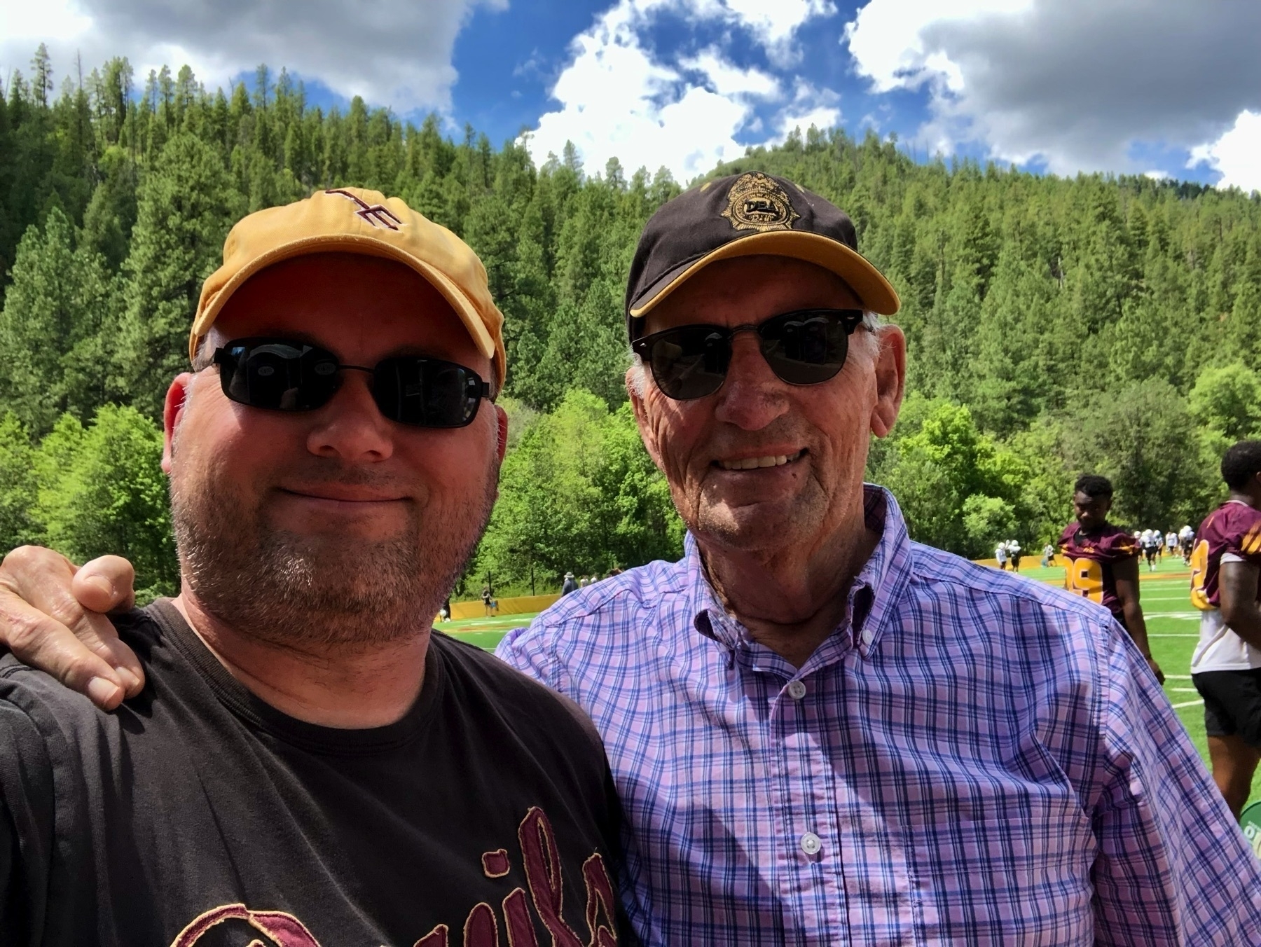 A selfie of my dad and I in front of the football field with pines in the background.