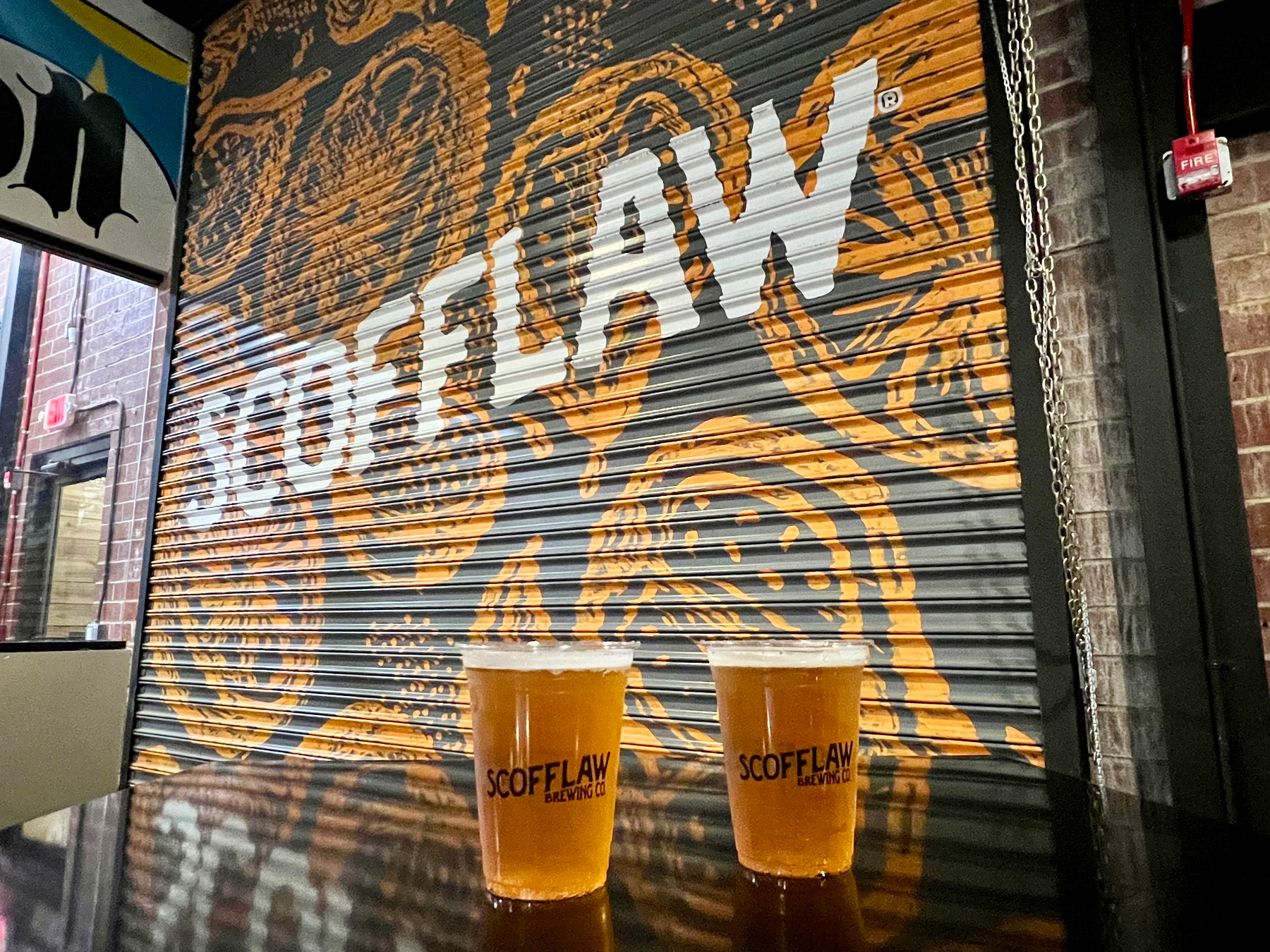 Two cups of beer sit on a table in front of a graffiti-style wall with the word SCOFFLAW.