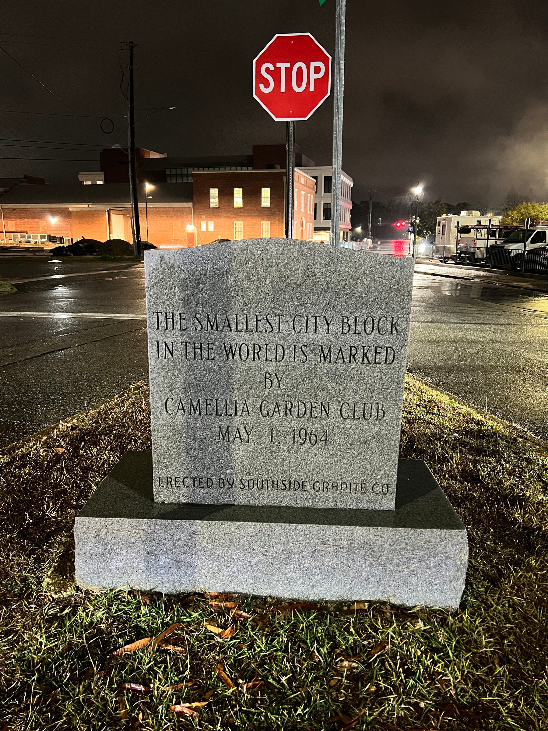 A granite marker commemorates the smallest city block in the world in front of a stop sign, with wet streets and buildings visible in the background.