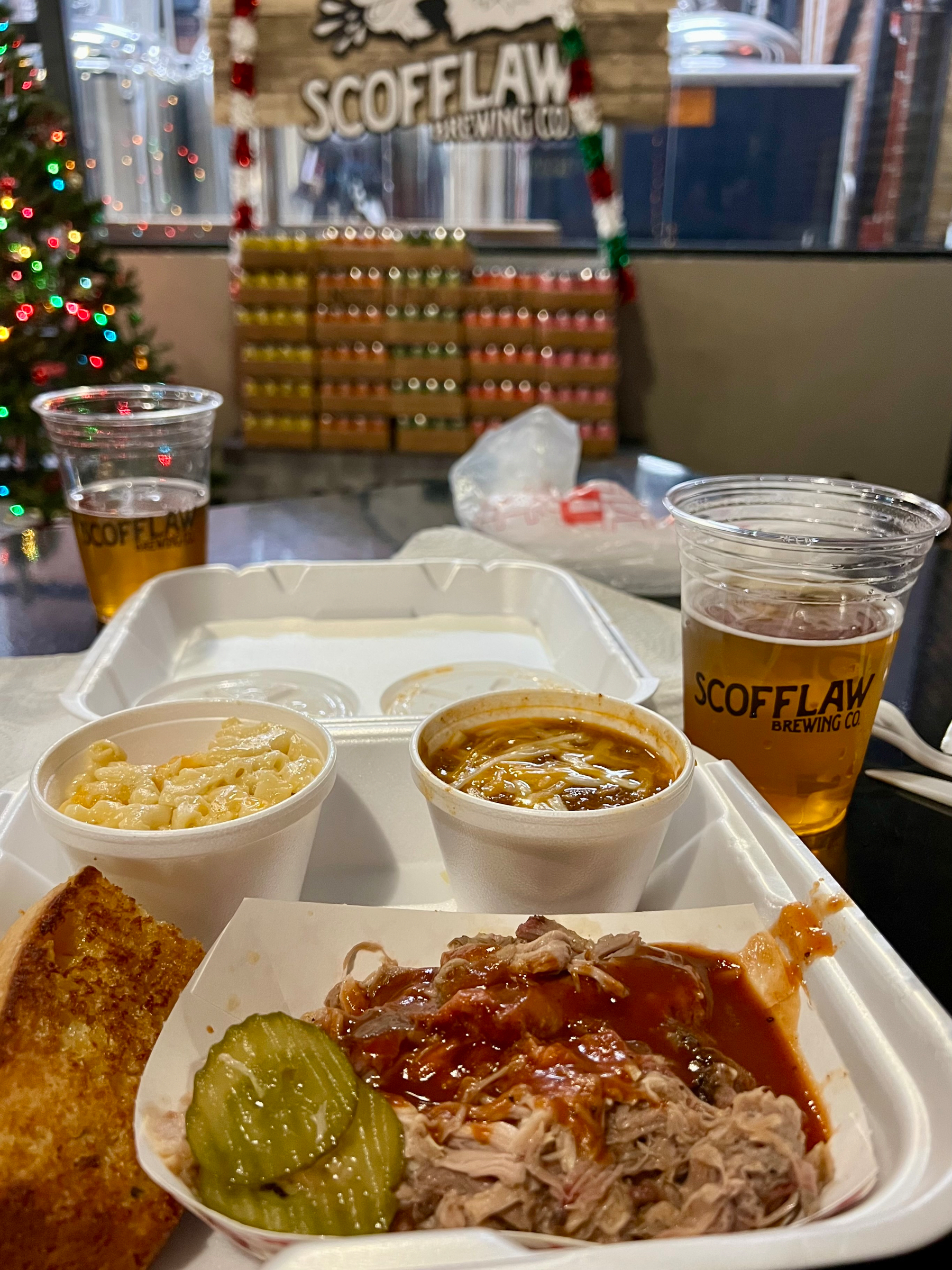 A meal consisting of pulled pork with barbecue sauce, pickles, macaroni and cheese, toasted bread, and drinks in plastic cups is set on a table at Scofflaw Brewing Co.