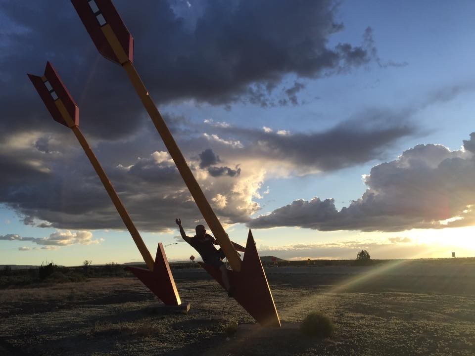 Auto-generated description: Two large arrow sculptures are planted in the ground beside a person striking a pose, set against a dramatic cloudy sky at sunset.