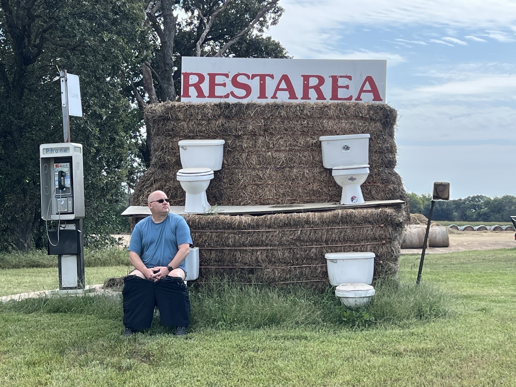 A person humorously poses near a makeshift "rest area" consisting of hay bales with toilets and a vintage phone booth nearby.