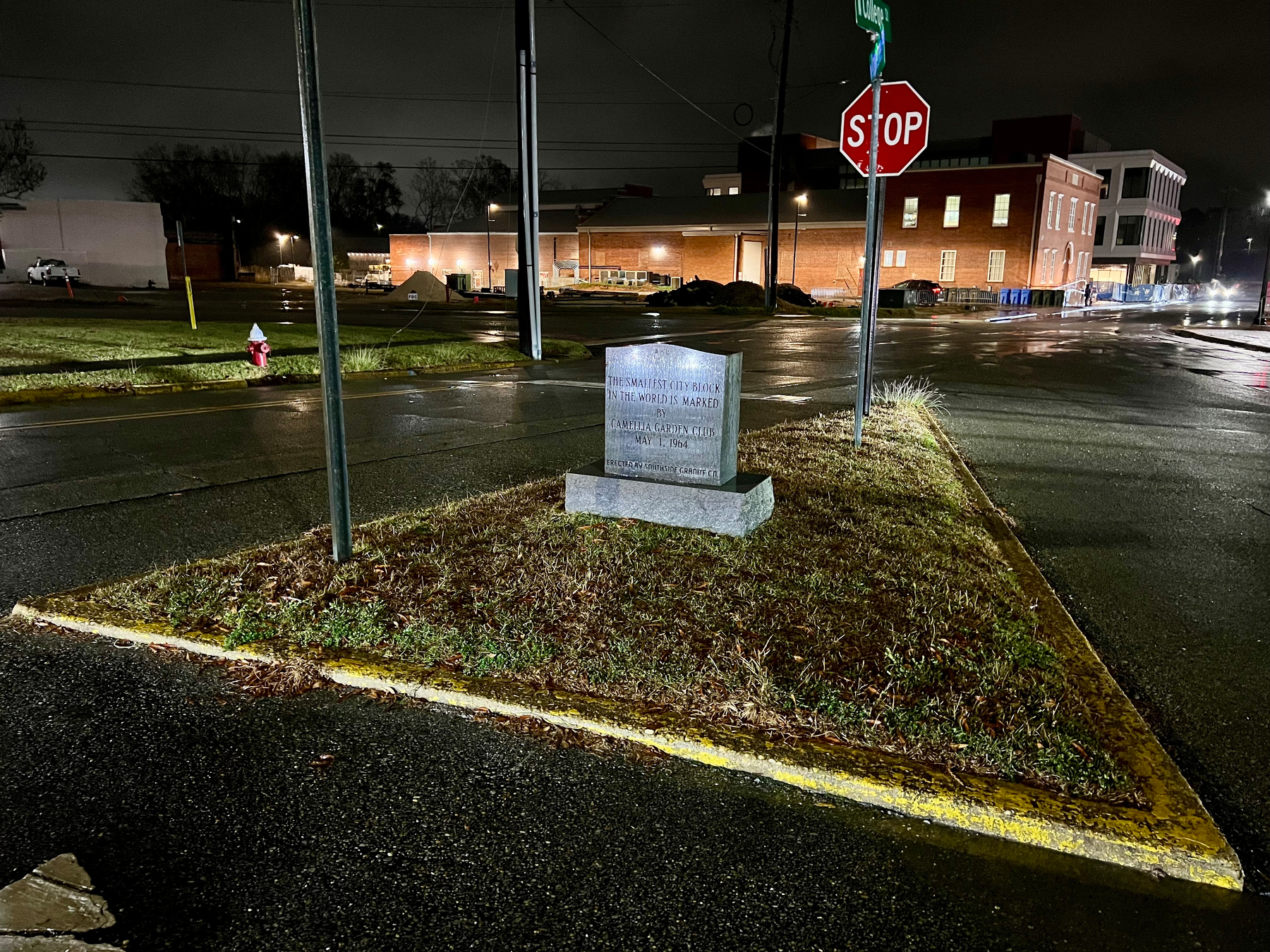 A dimly lit street corner features a triangular patch of grass with a stone marker displaying inscriptions, set against a backdrop of buildings and a wet road, accented by a stop sign.