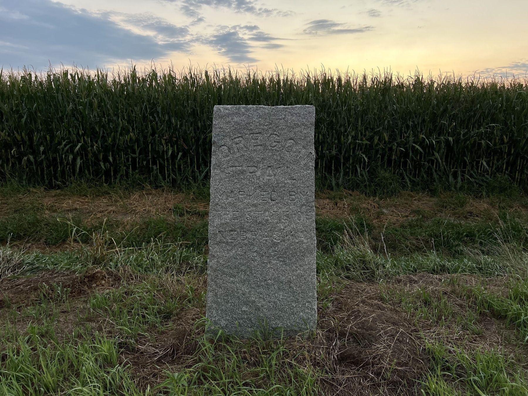 A stone monument stands in front of a cornfield at sunset.