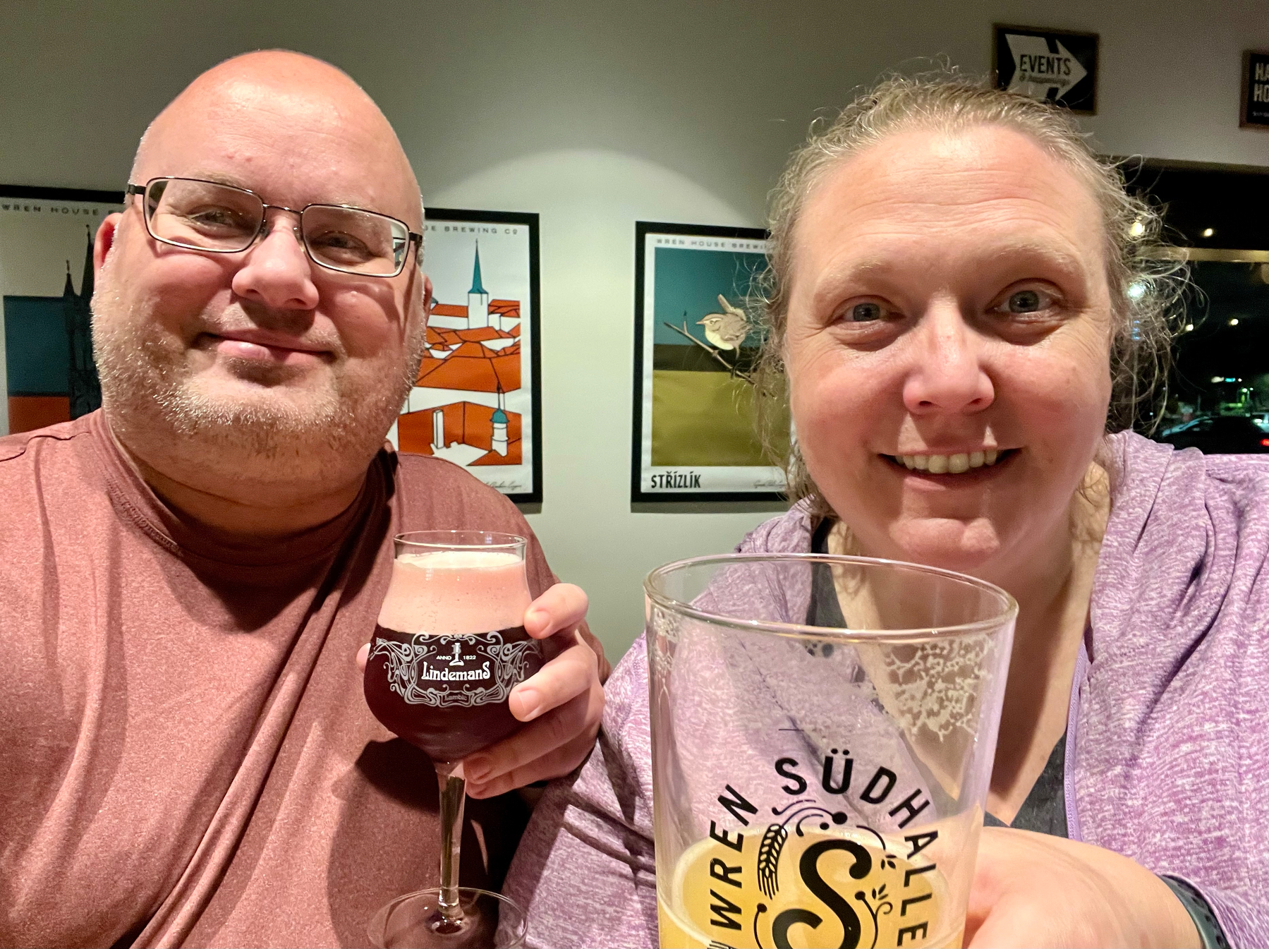 Two people are smiling and holding glasses of dark beer with artwork on the wall behind them.