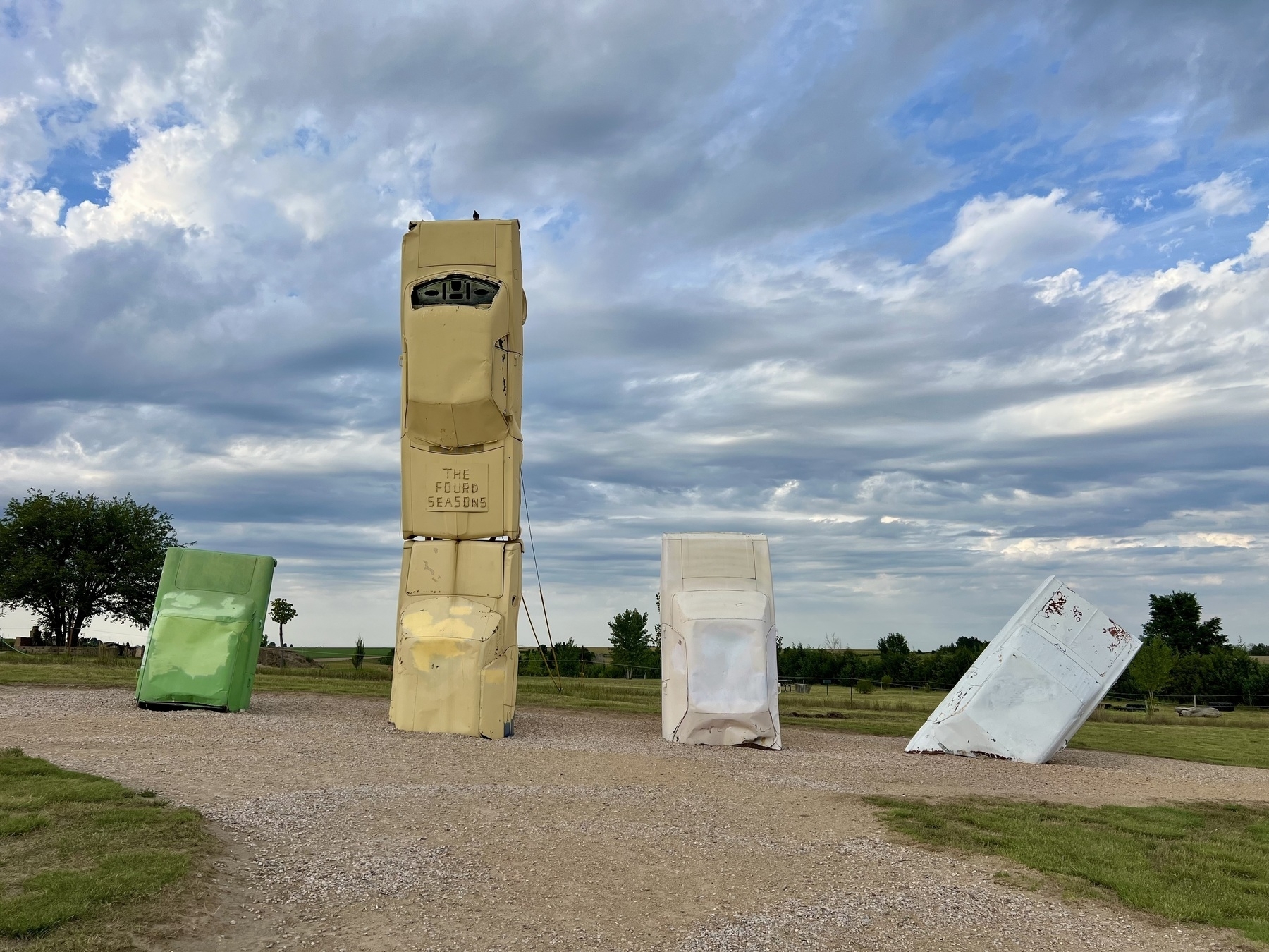 Several cars are artistically arranged standing nose-down in the ground with one car balanced on top, set against a landscape with a cloudy sky.