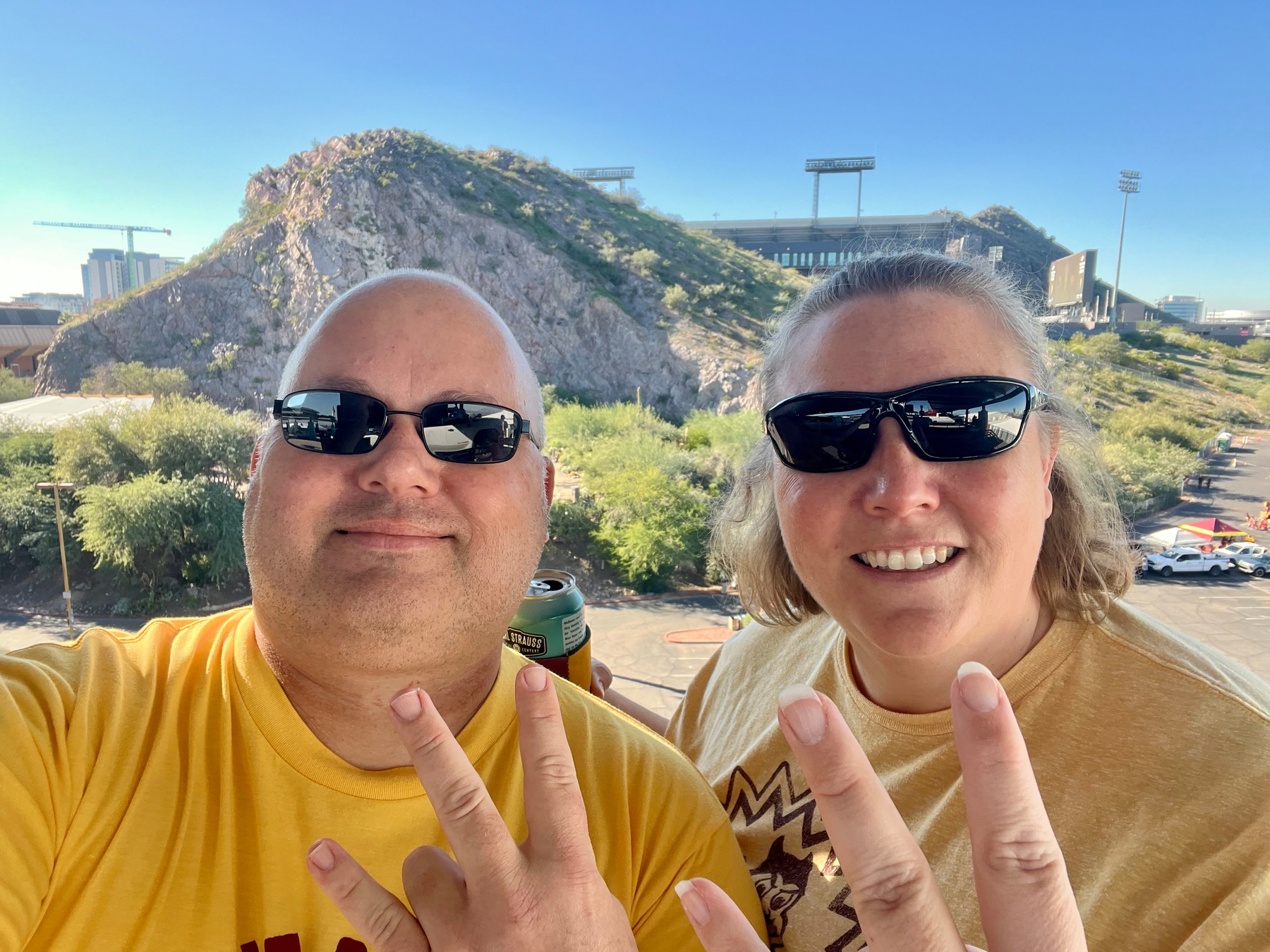 Two people wearing sunglasses are posing with raised pitchforks, with a rocky hill and Sun Devil Stadium in the background.