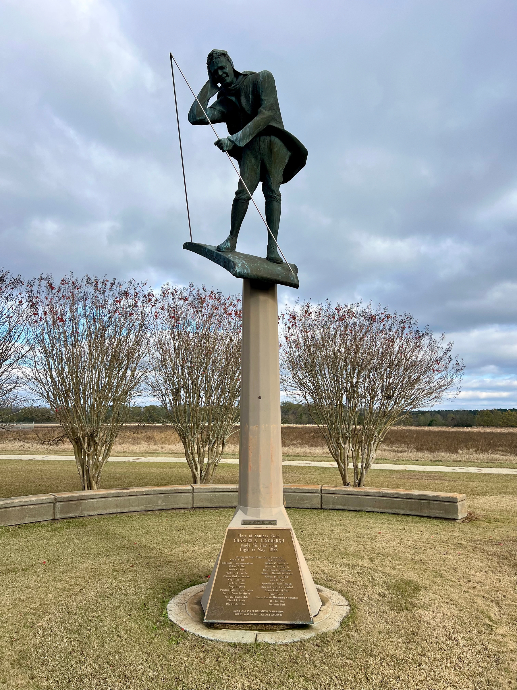 A statue of a man with a fishing rod is mounted on a tall pedestal surrounded by landscaped grass and trees.