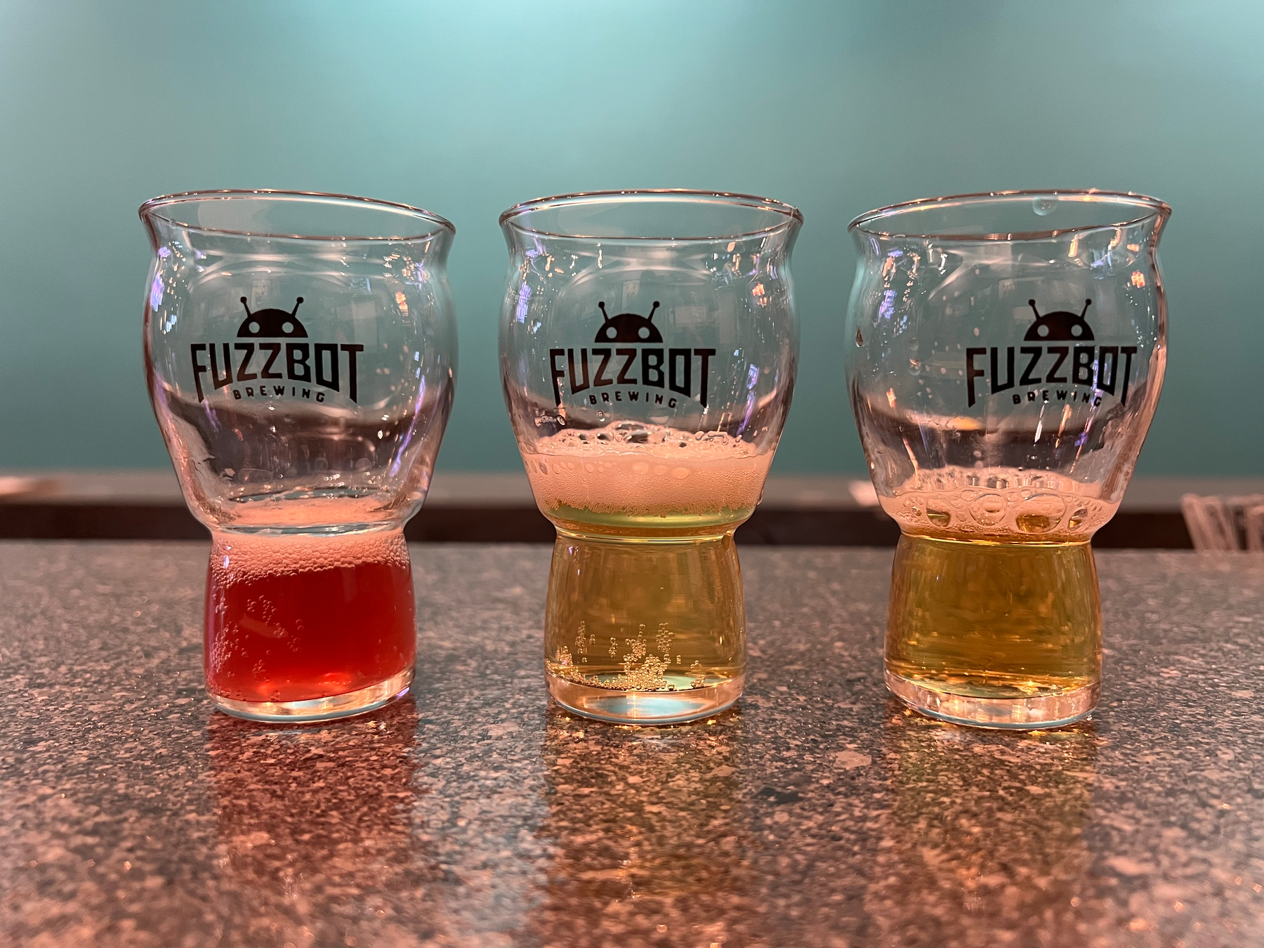 Three small glasses with varying amounts of different colored liquids are arranged on a speckled countertop, each labeled with a FUZZBOT BREWING logo.