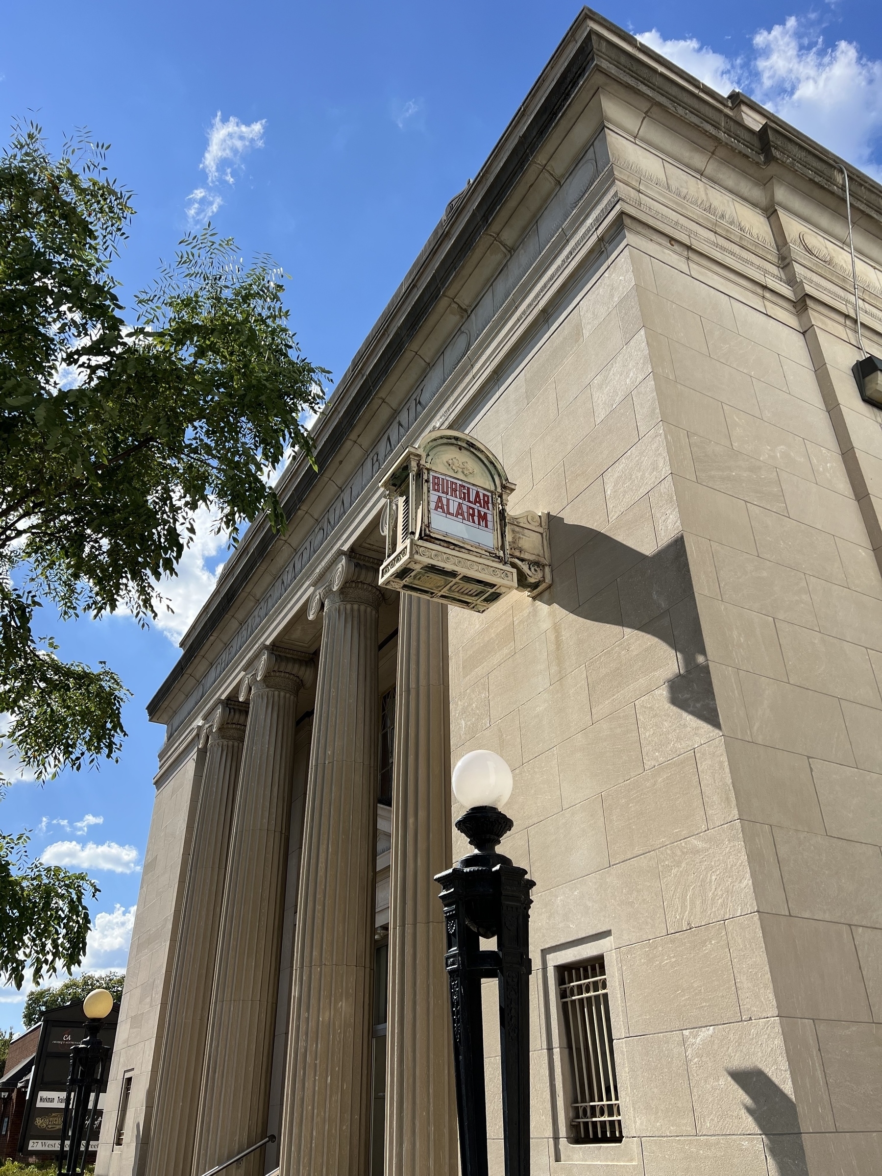 A classical-style building with large columns and a "BURGLAR ALARM" sign near the entrance is set against a backdrop of a blue sky with scattered clouds and a leafy tree.