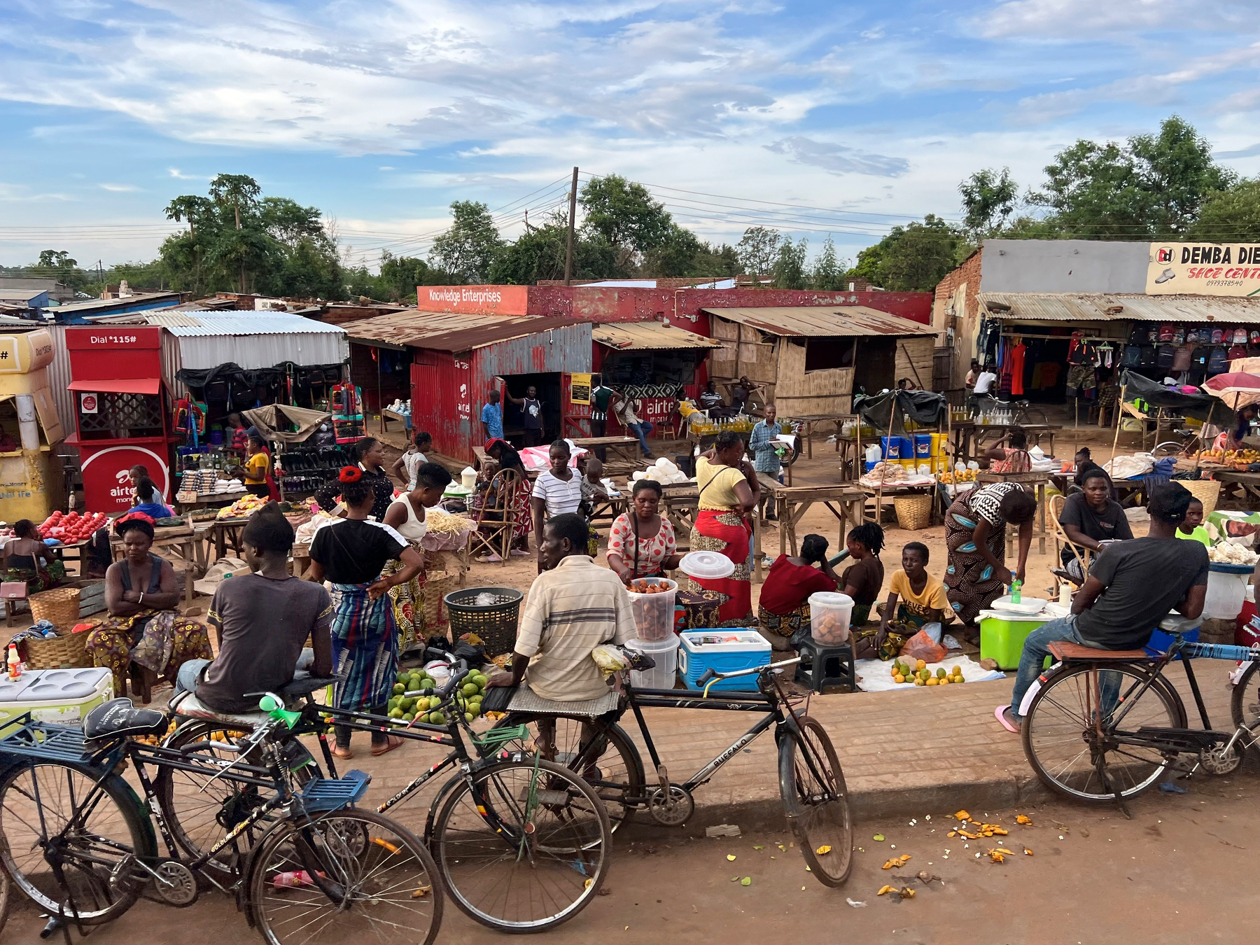 A bustling outdoor market scene features vendors selling goods and people engaging with various stalls, with bicycles parked in the foreground.