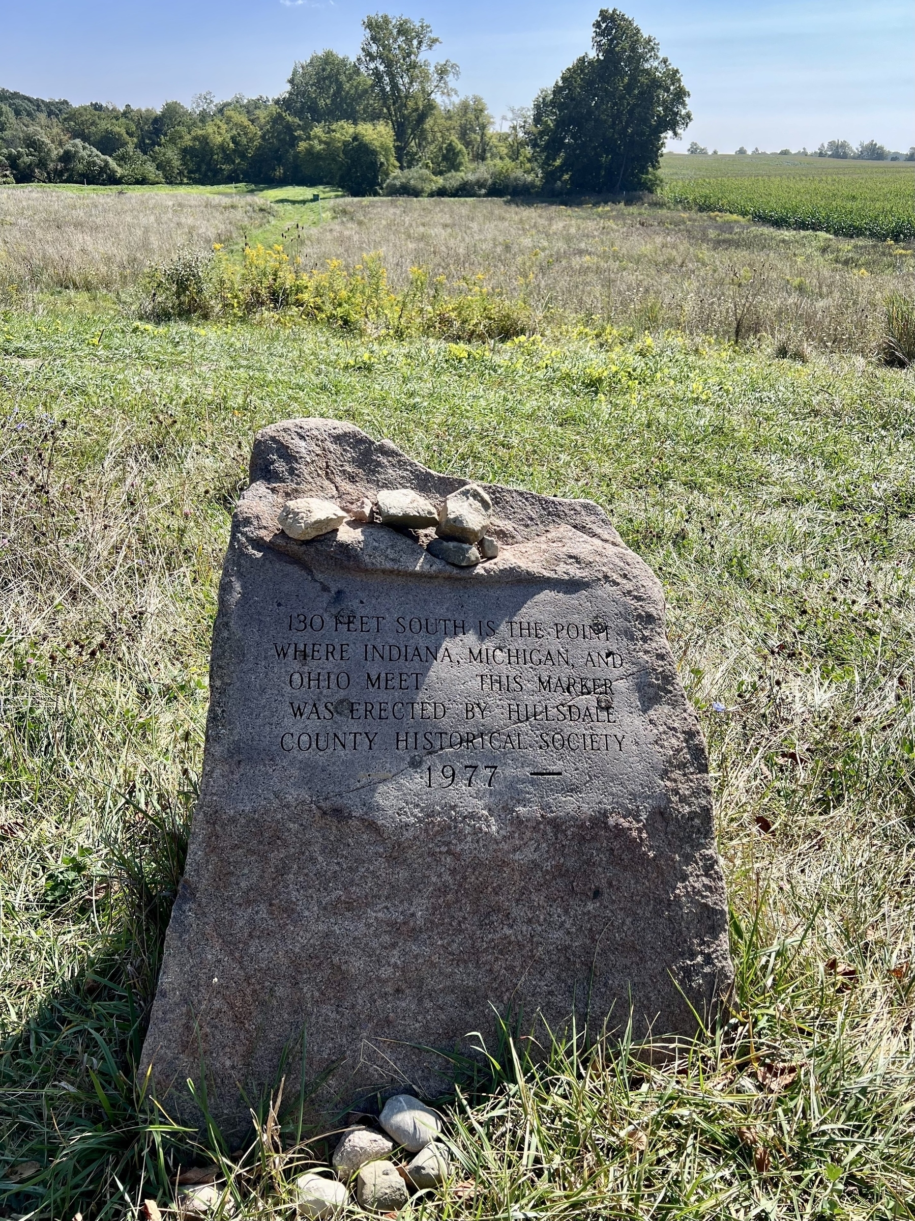 A commemorative stone marker in a grassy field indicates the nearby junction of Indiana, Michigan, and Ohio, marked by the Hillsdale County Historical Society in 1977.