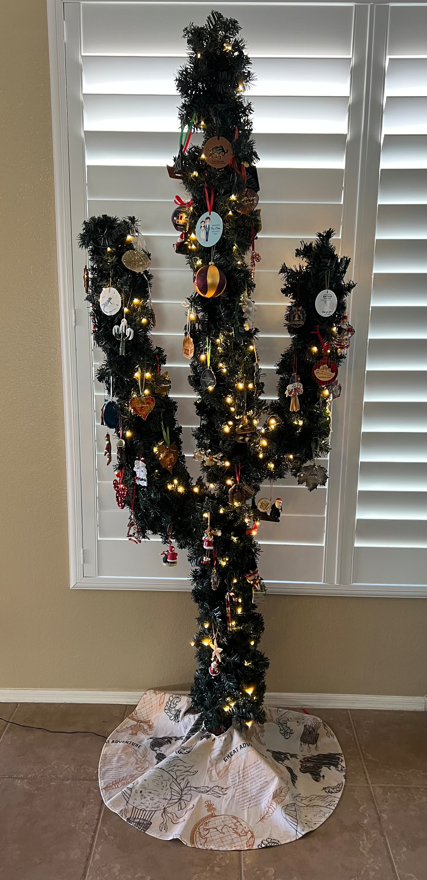 A cactus-shaped Christmas tree, adorned with lights and ornaments, stands in front of a window with shutters.