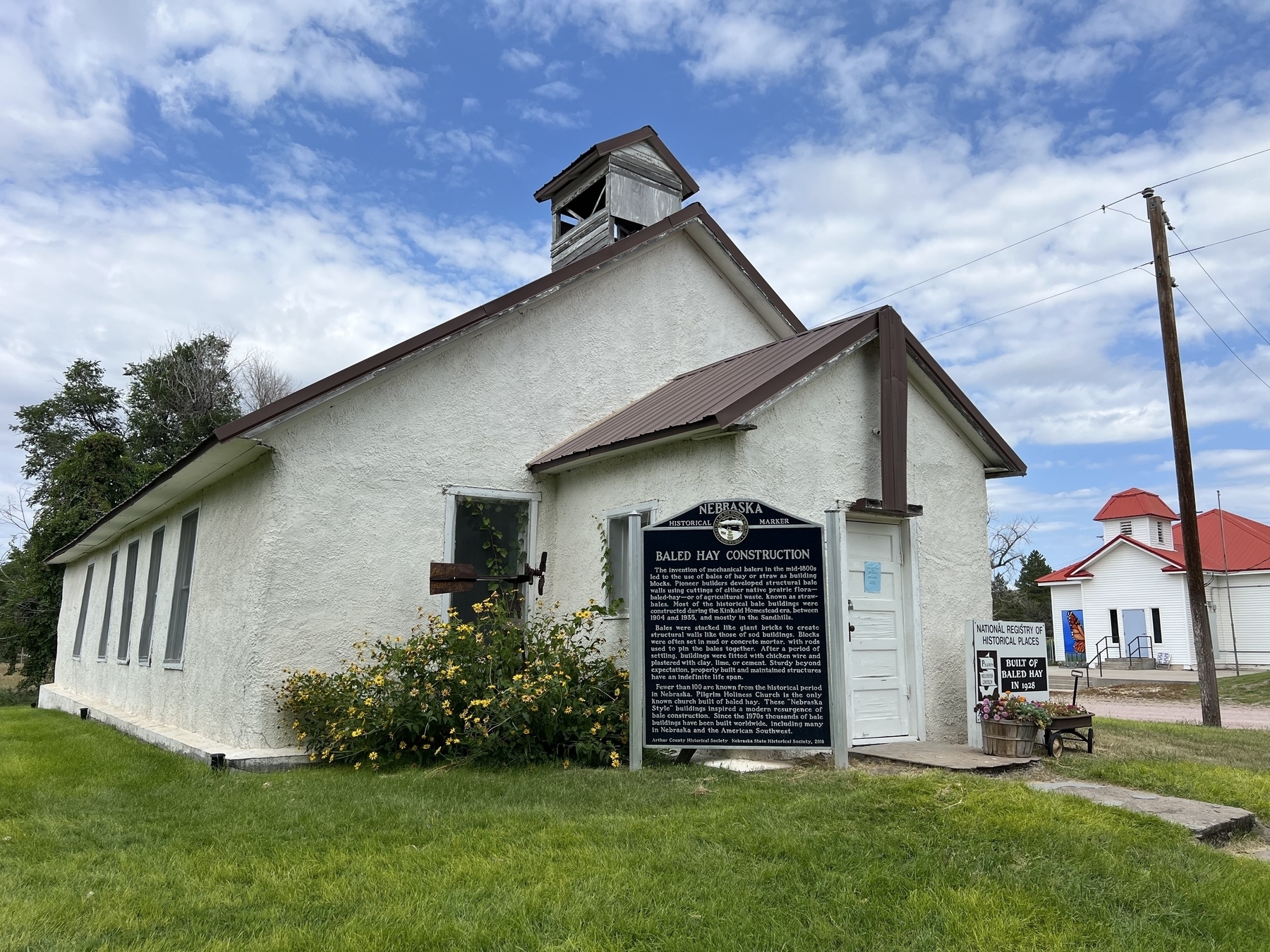 A historical white building with a sign detailing its construction and significance stands under a blue sky with scattered clouds.