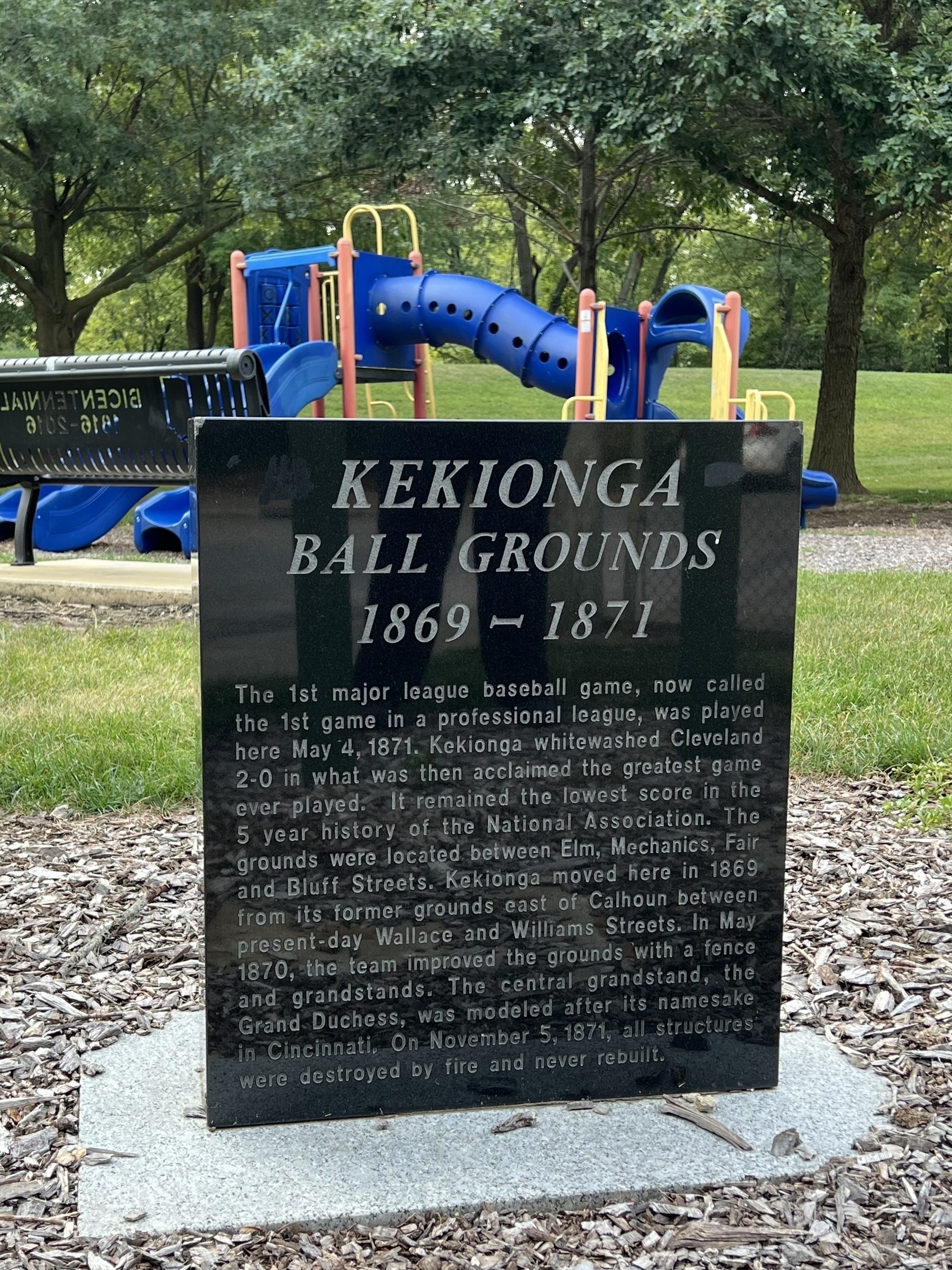 A dark monument surrounded by grass and trees commemorates the Kekionga Ball Grounds, detailing historical dates and events from 1869 to 1871.