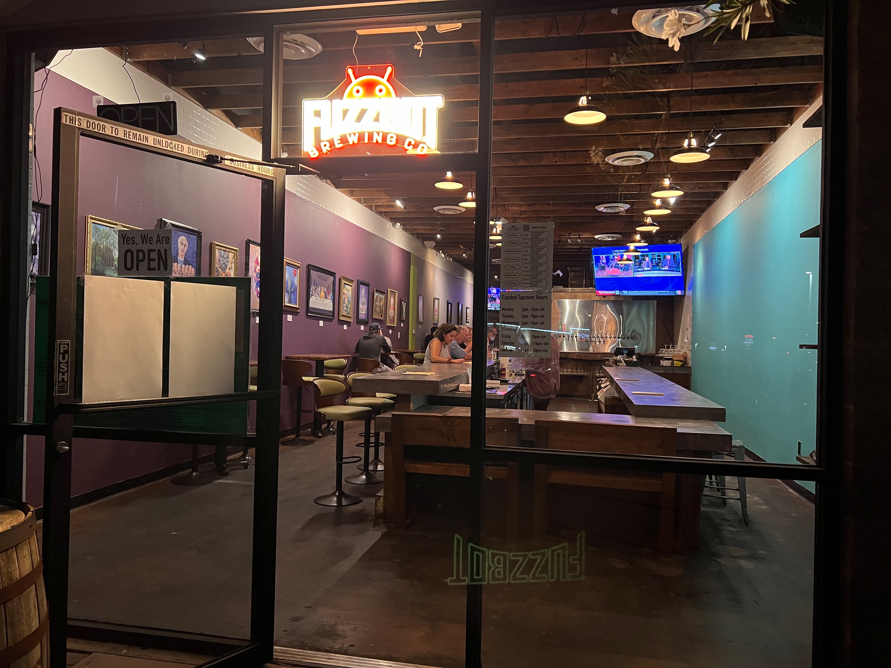 A cozy, dimly lit bar features colorful artwork on the walls, a few patrons seated at the counter, and a neon sign reading FUZZBOT BREWING CO. hanging in the window.