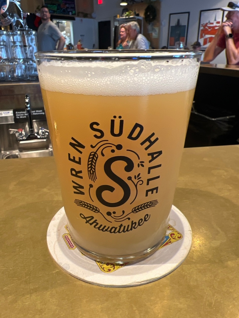 A large glass of beer with a foamy head is on a coaster at a bar, featuring the text Wren Südhalle Ahwatukee on its side.