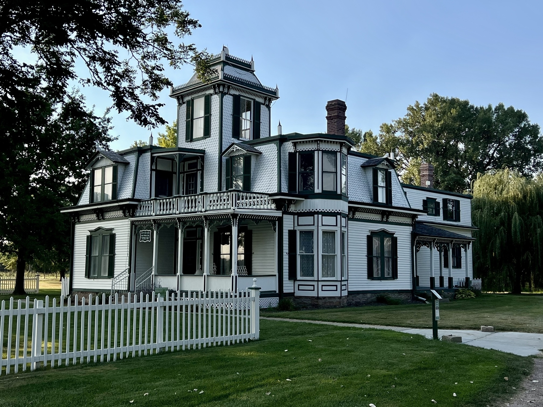 A large, historic Victorian-style house with intricate detailing is surrounded by a white picket fence and lush greenery.