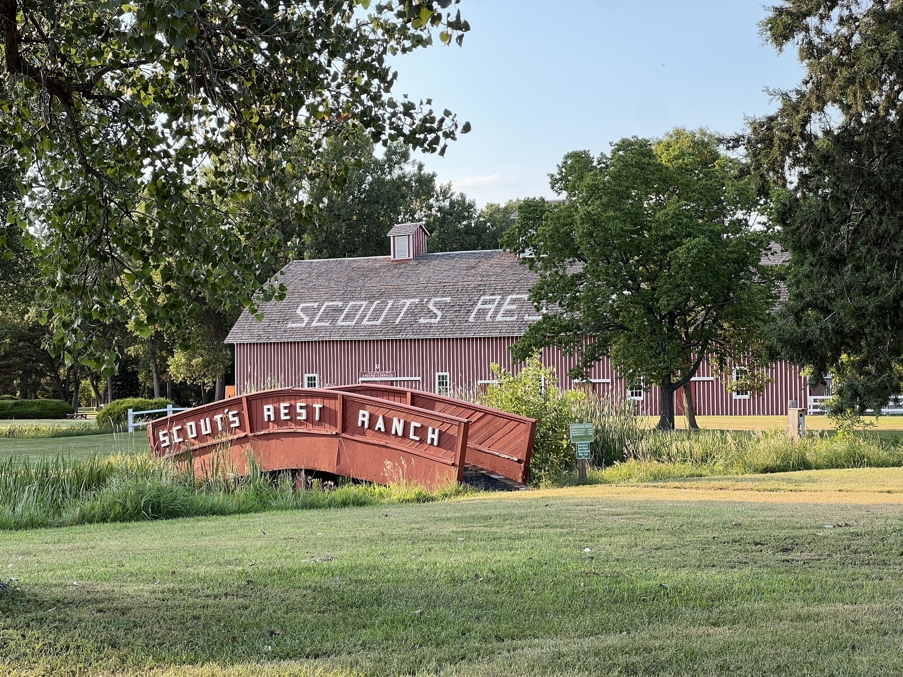 A red barn with the words "Scouts Rest Ranch" on the roof is situated behind a red footbridge with the same inscription, surrounded by trees and a grassy area.