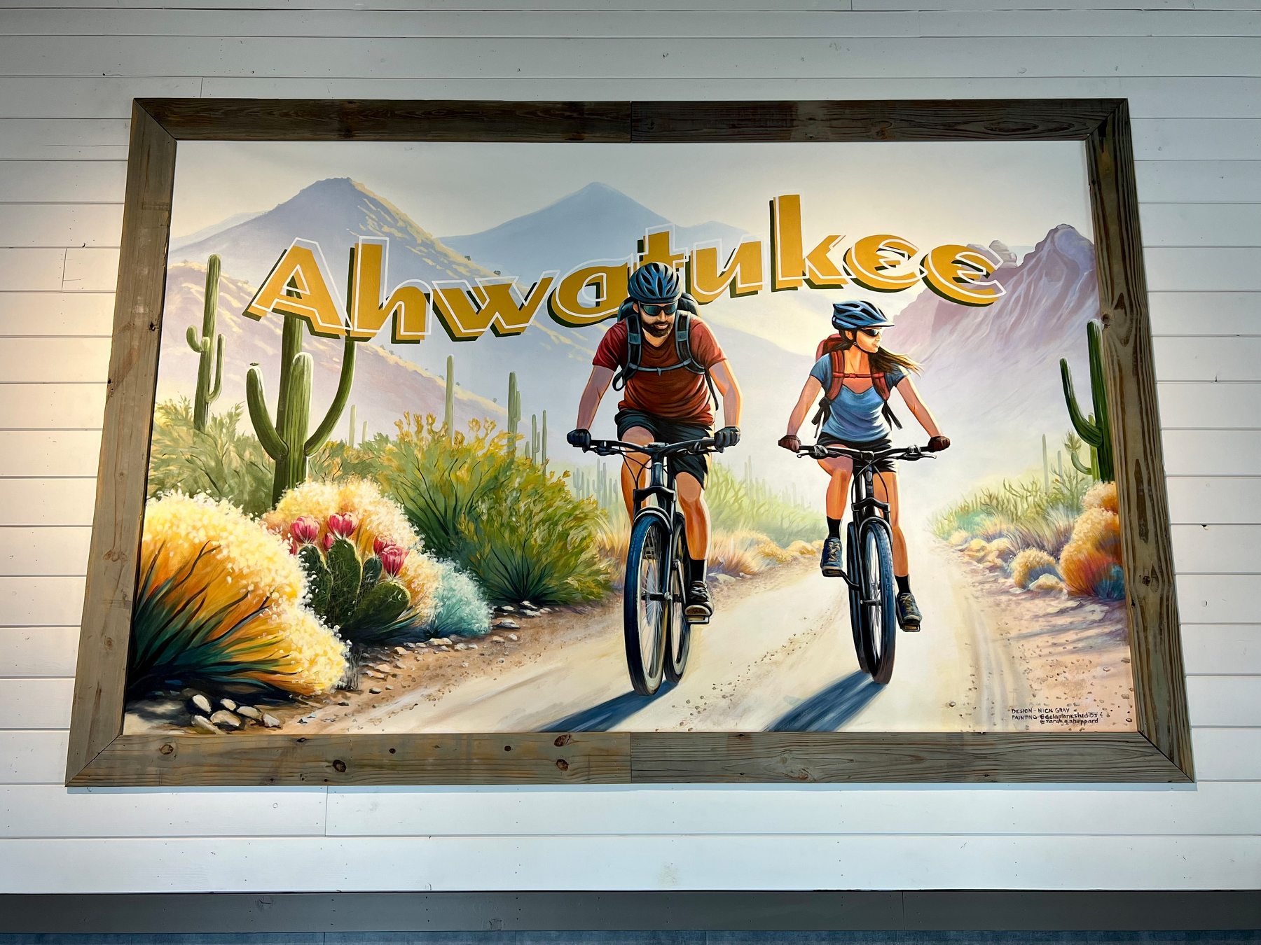A mural depicts two cyclists riding on a desert trail surrounded by cacti and mountains, with the word Ahwatukee prominently displayed above.