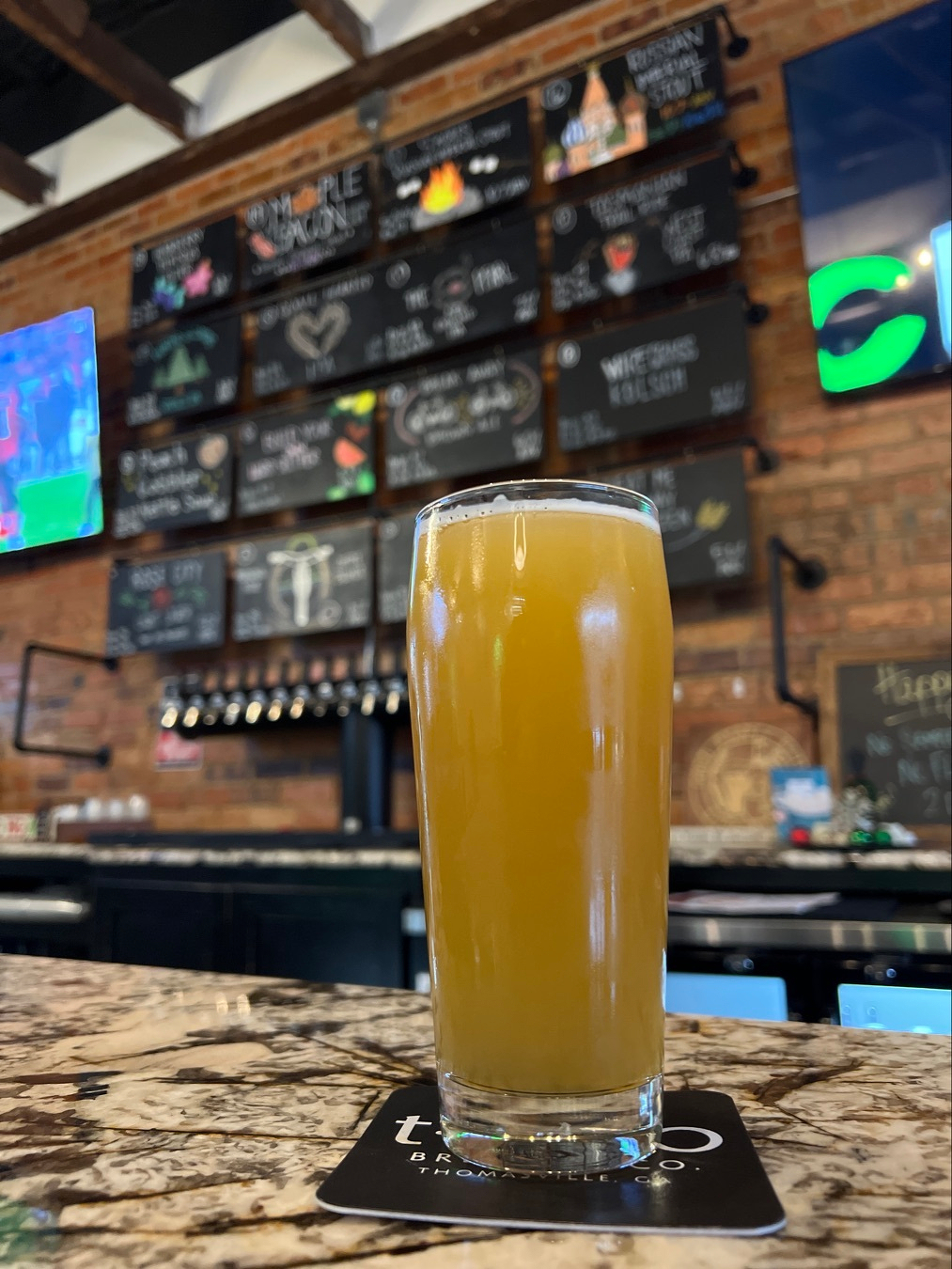 A glass of beer sits on a bar counter, with a background of colorful chalkboard menus.