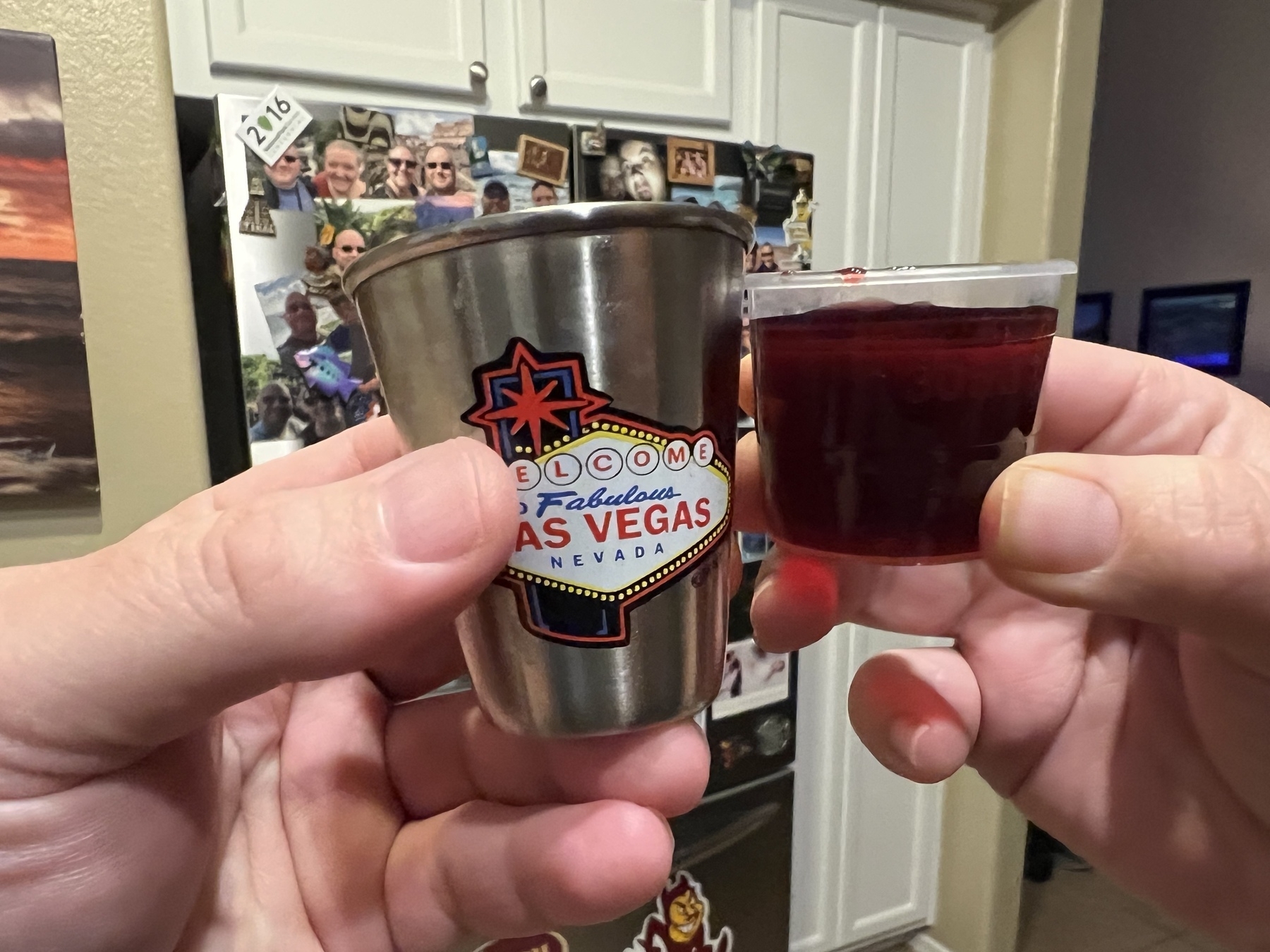Two hands are holding a Las Vegas shot glass and a small cup filled with a red liquid in a kitchen.