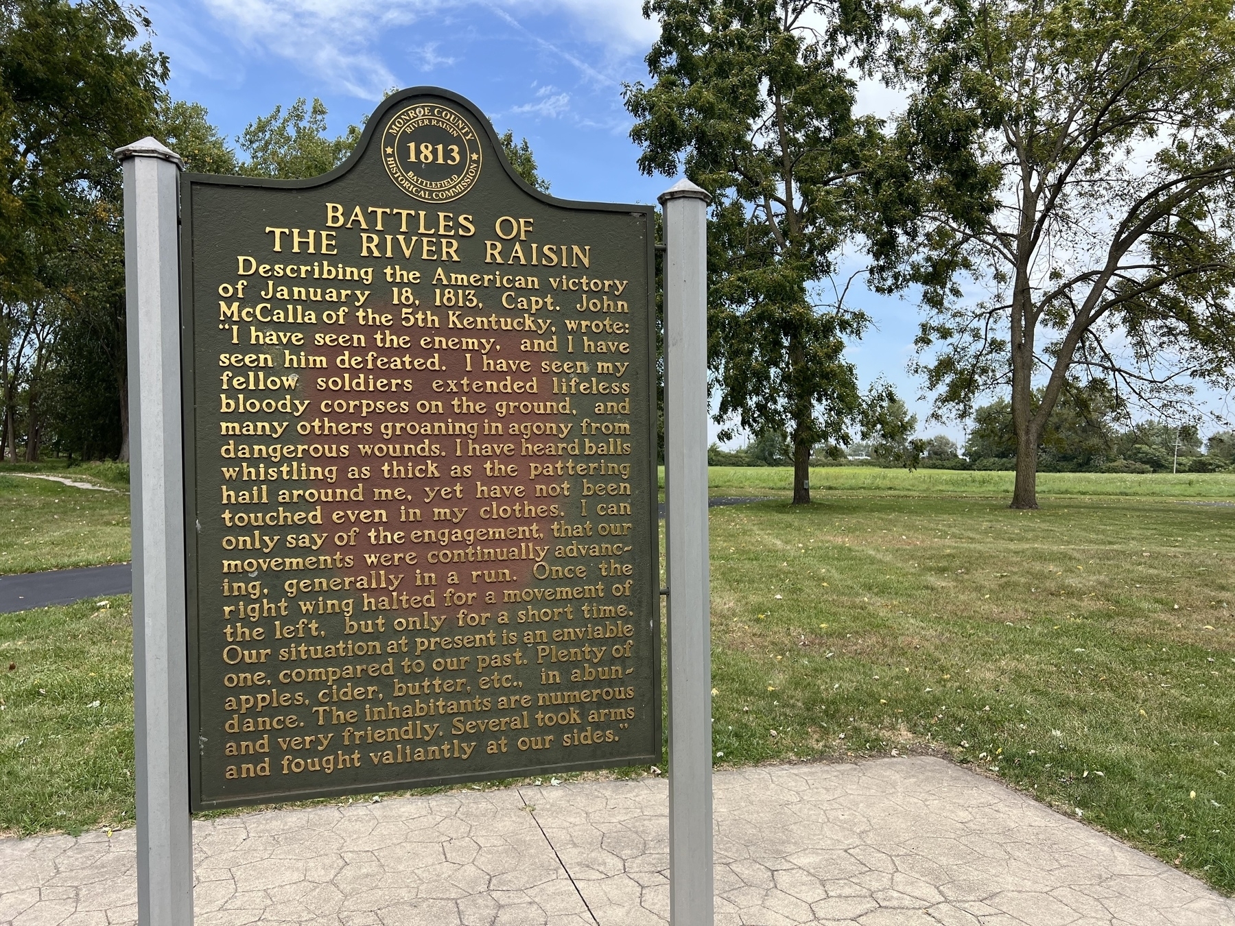A historical marker detailing the Battles of the River Raisin is set against a background of green grass, trees, and a partly cloudy sky.