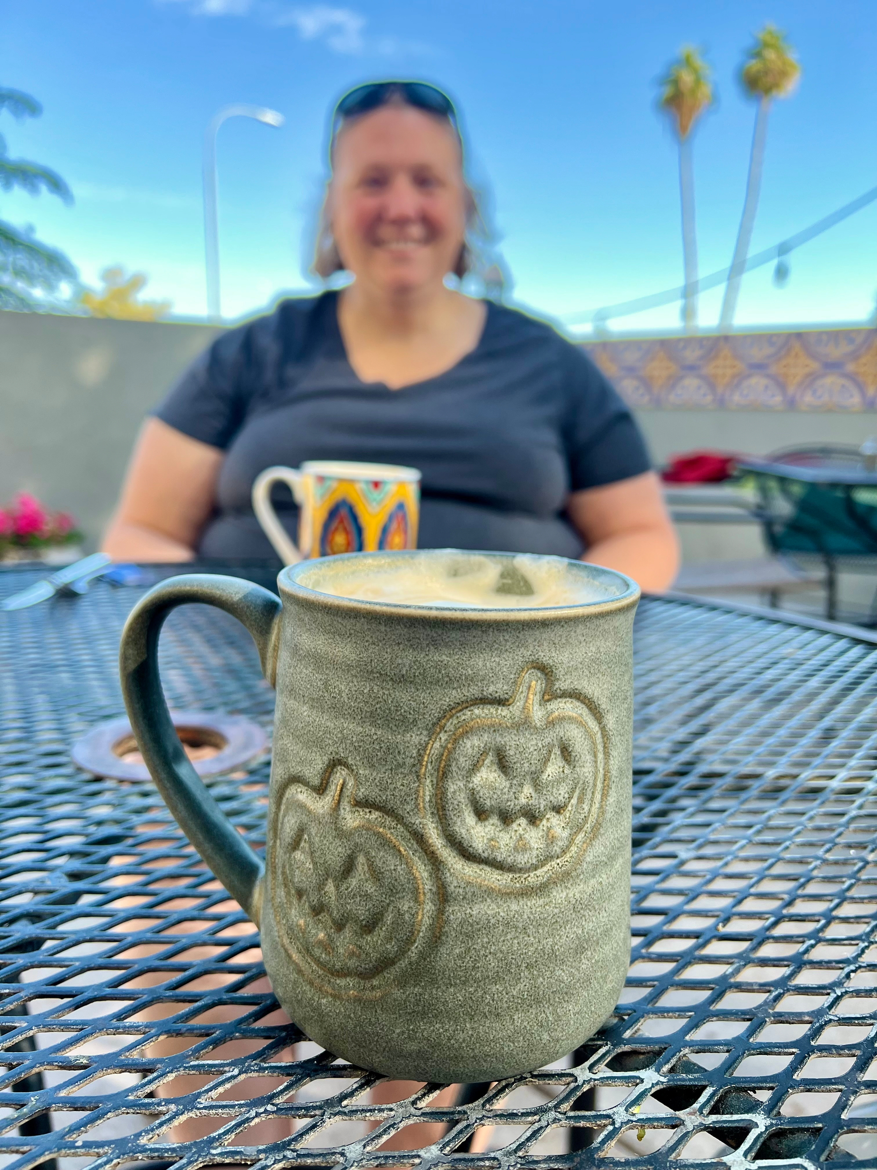 A ceramic mug with pumpkin designs is on a table, with a person sitting and smiling in the background.