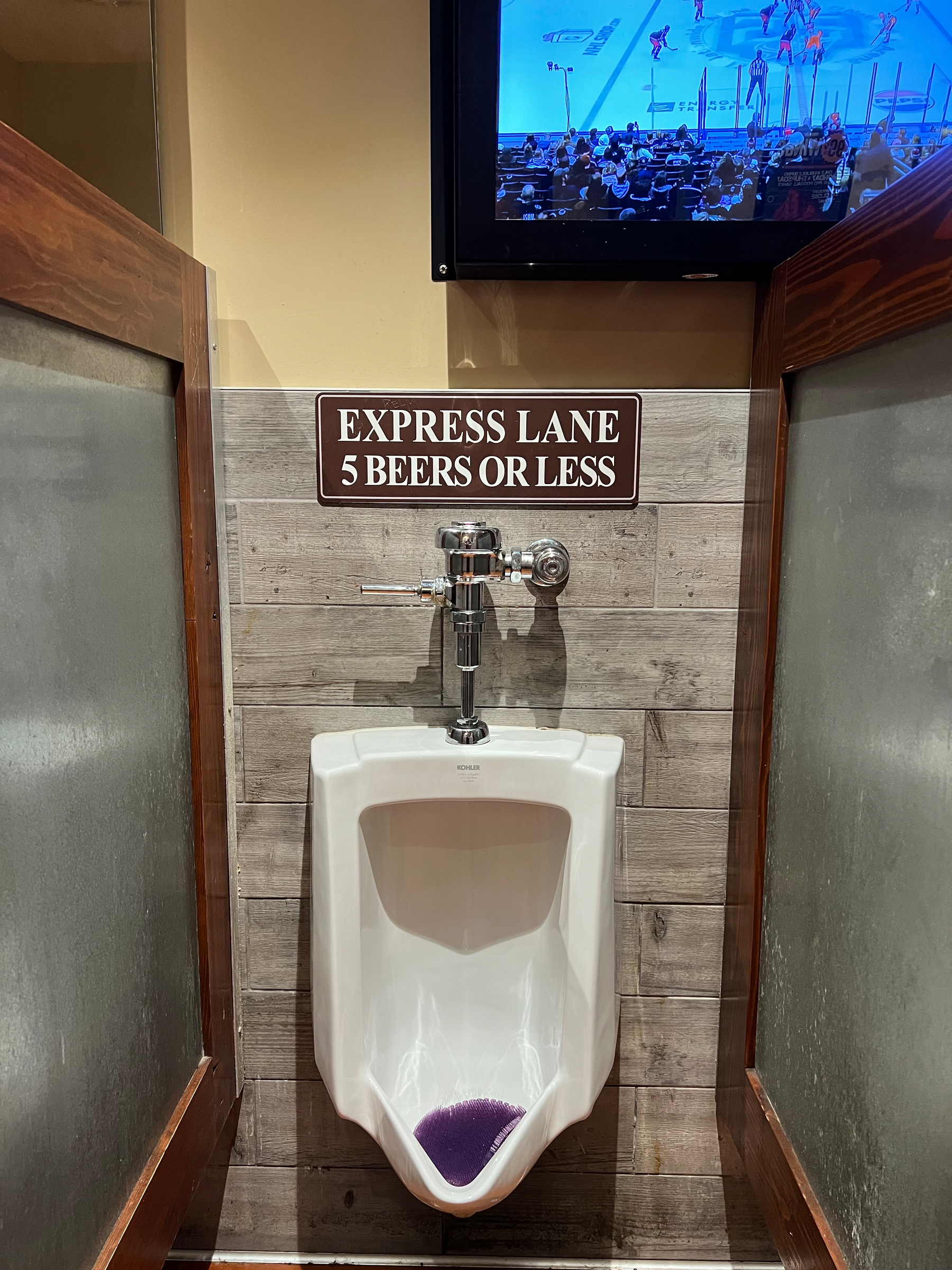 A urinal with a humorous sign above it reading EXPRESS LANE 5 BEERS OR LESS and a TV screen showing a sports game.