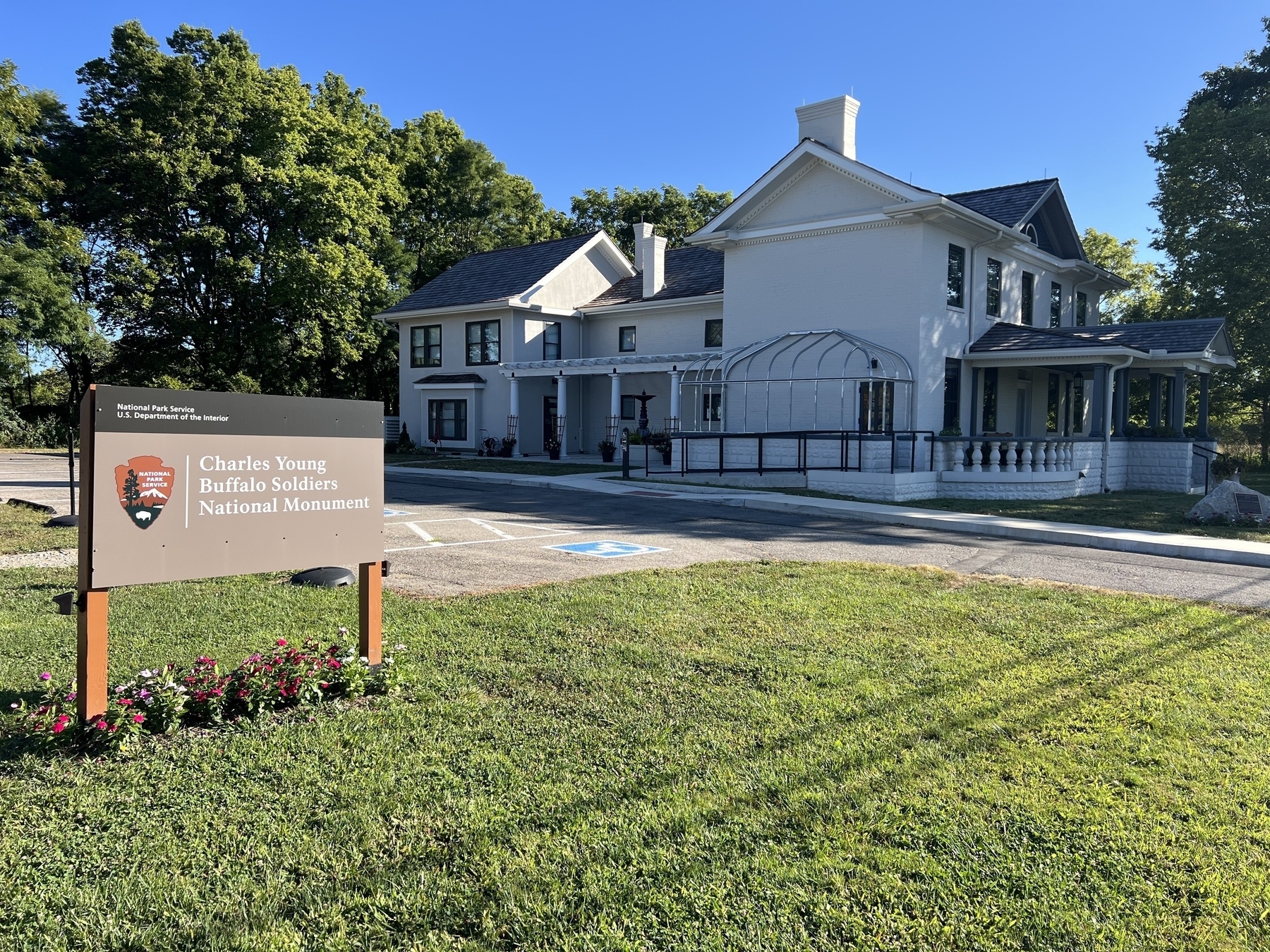 A white historic building is situated beside a sign for the Charles Young Buffalo Soldiers National Monument, surrounded by greenery and clear blue skies.