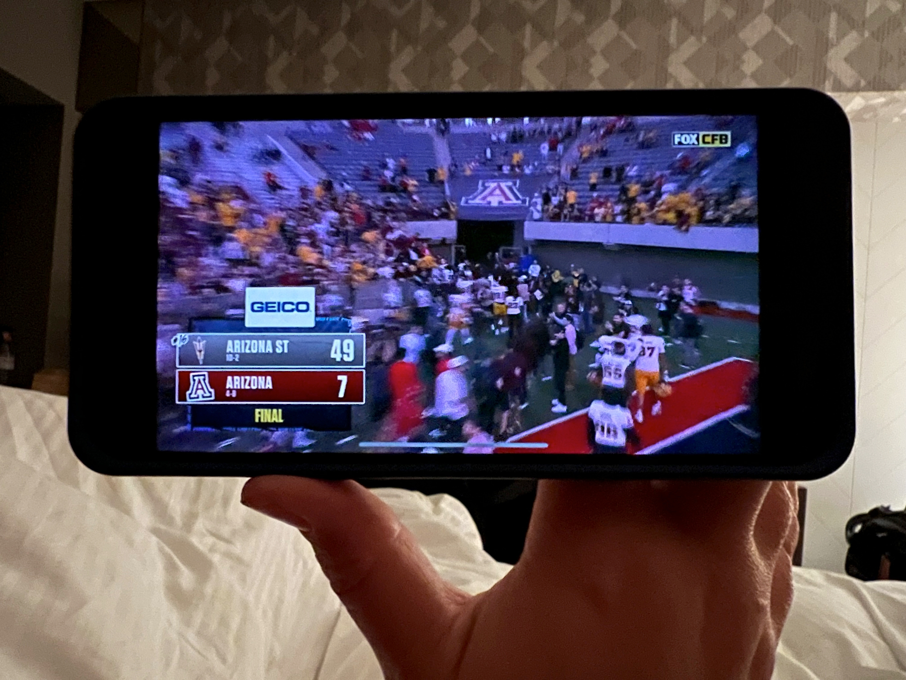 A person is holding a smartphone displaying a football game with a final score of Arizona State 49 and Arizona 7.