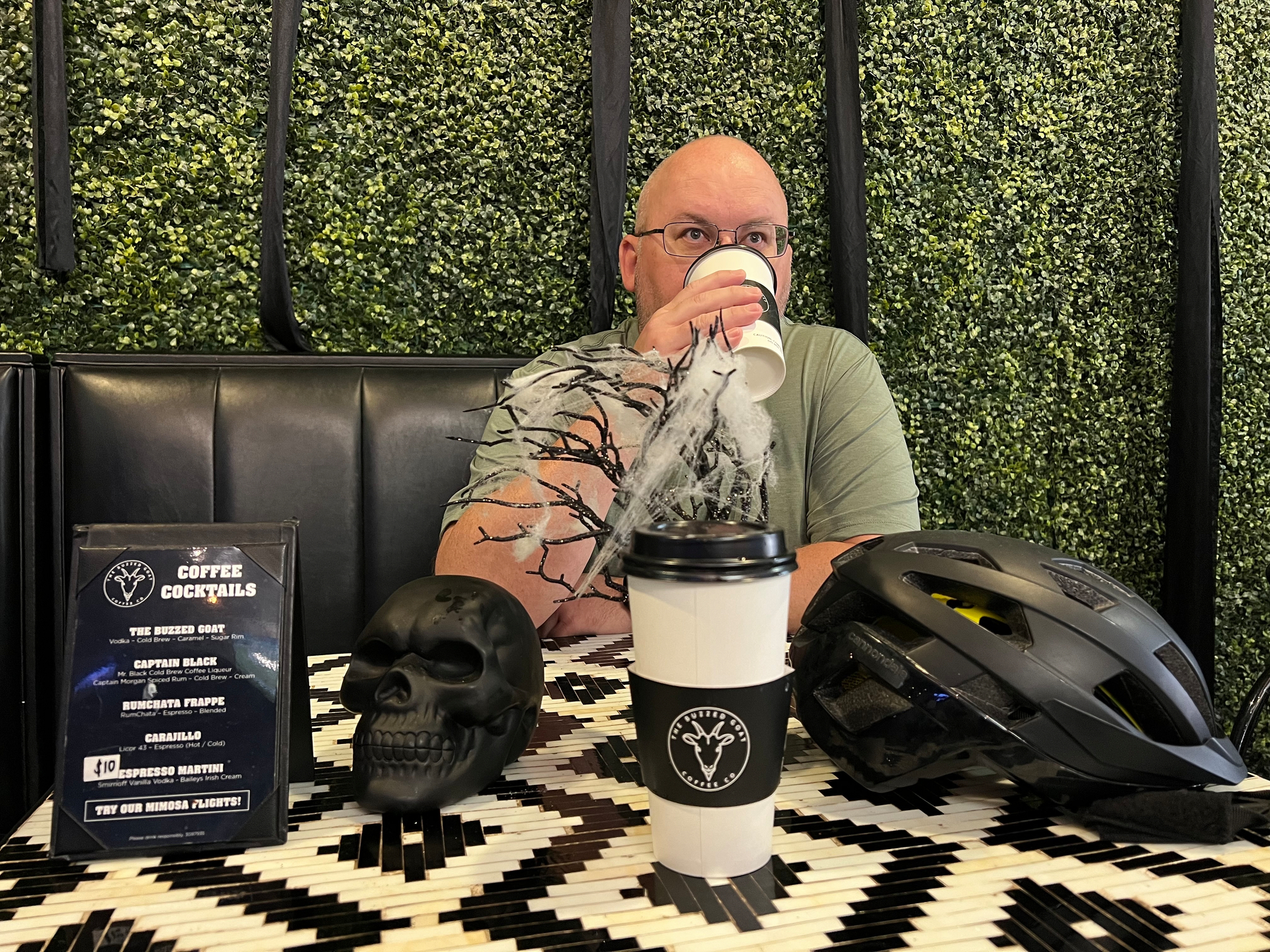 A man sipping a coffee sits at a table with Halloween-themed decor, including a skull and a black helmet, in front of a backdrop of green foliage, while participating in Coffeeneuring.
