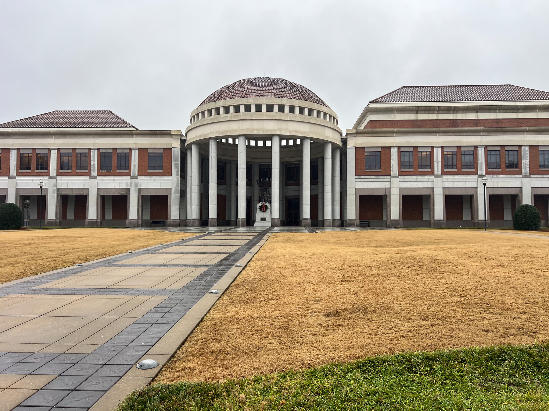A large building with a dome and columns stands at the end of a paved walkway, surrounded by a grassy area.