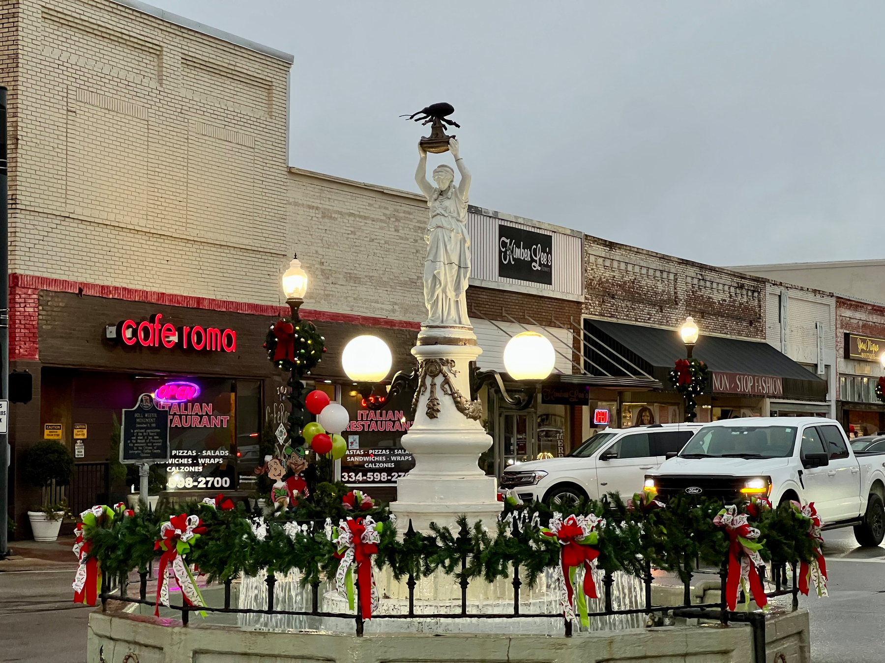 A MOTHAFUCKIN BOLL WEEVIL statue surrounded by festive decorations stands in the center of a small, decorated town square with various shops in the background.