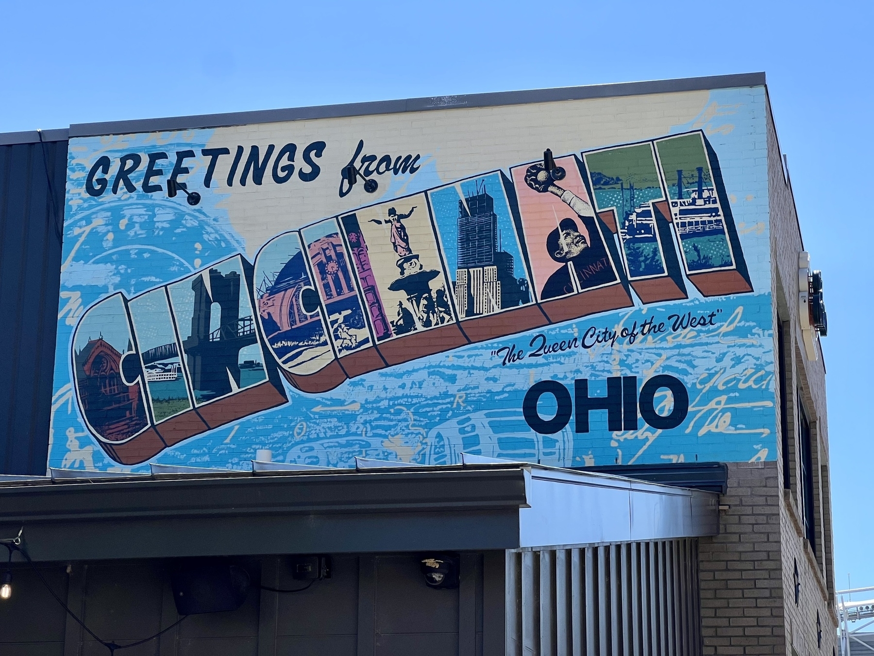 A colorful mural on a building features the phrase "Greetings from Cincinnati, Ohio" with local landmarks illustrated within the letters.