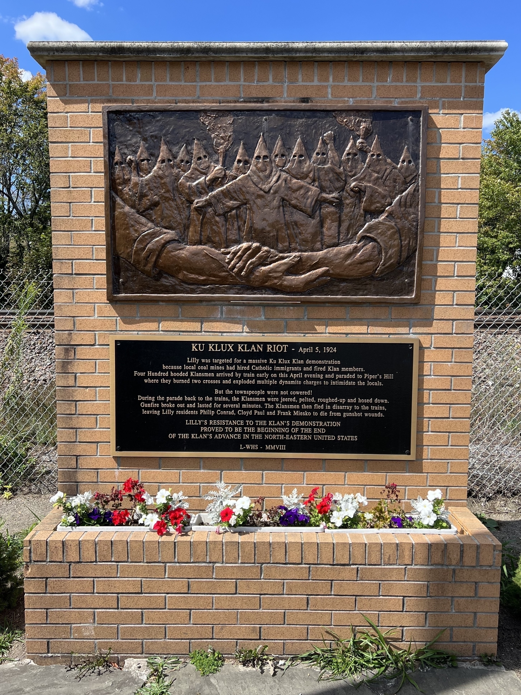 A brick monument features a relief of hooded figures with a plaque describing the Ku Klux Klan riot of April 1, 1934, surrounded by colorful flowers.