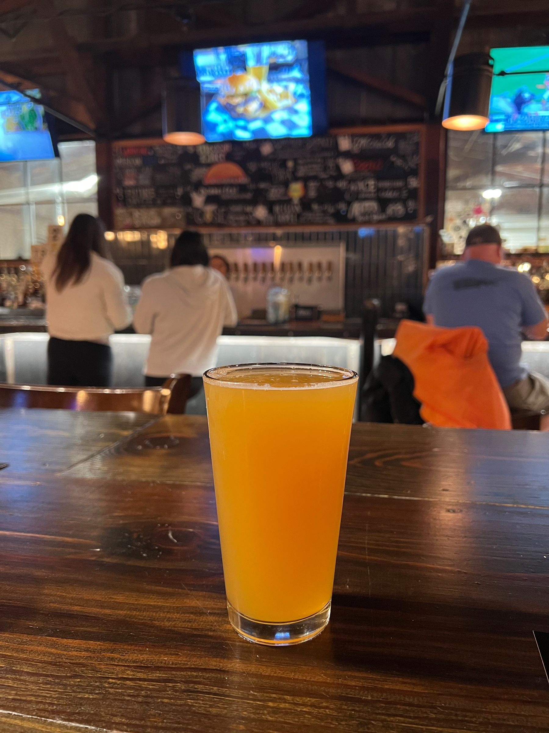 A pint of beer is placed on a wooden bar counter in a busy pub with patrons and televisions in the background.