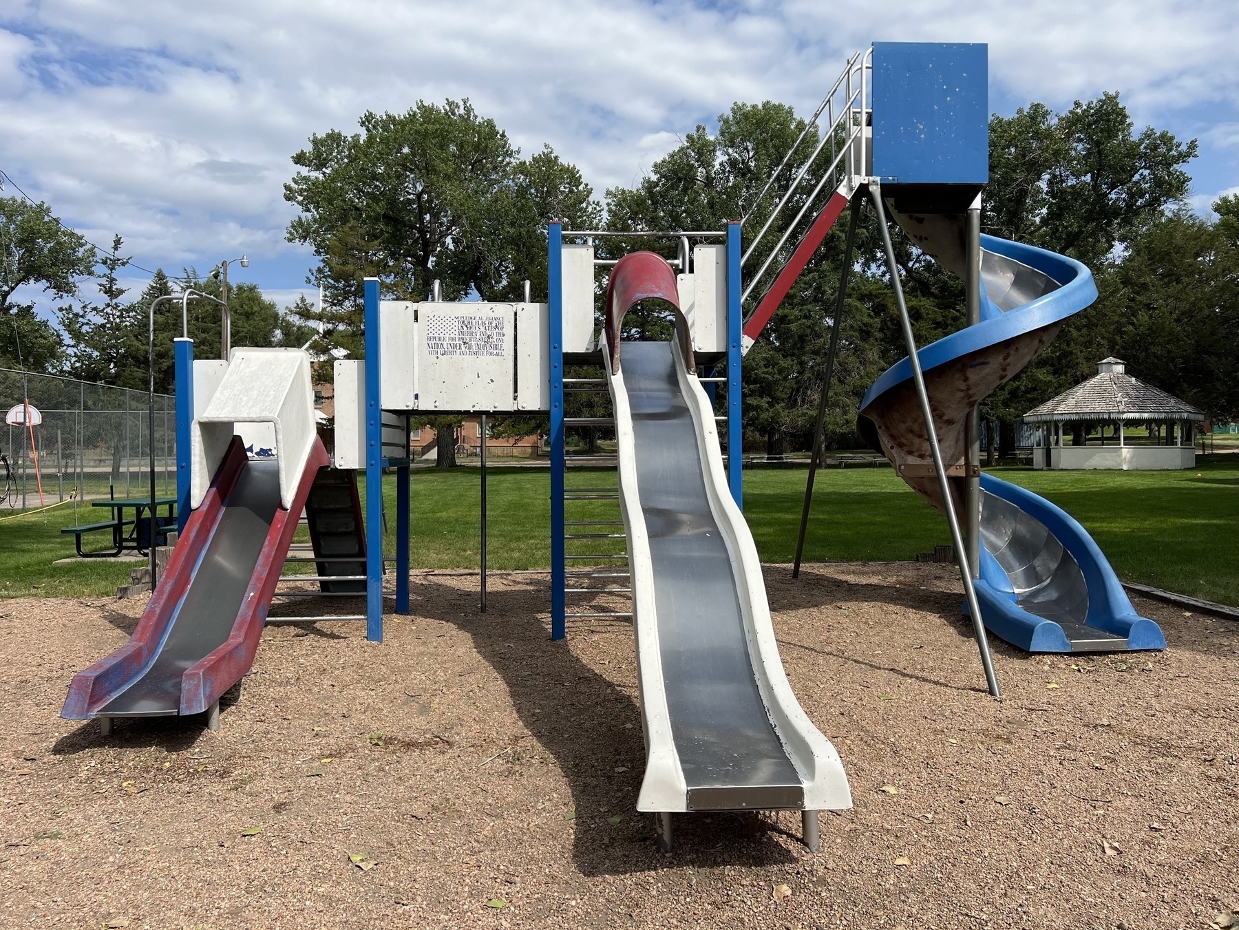 A playground features multiple slides and climbing structures on a gravel surface, surrounded by trees and open grassy areas.