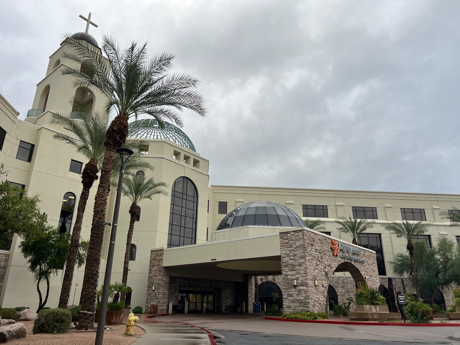 A large hospital building with a prominent church-like dome and cross, surrounded by palm trees, is seen under a cloudy sky.