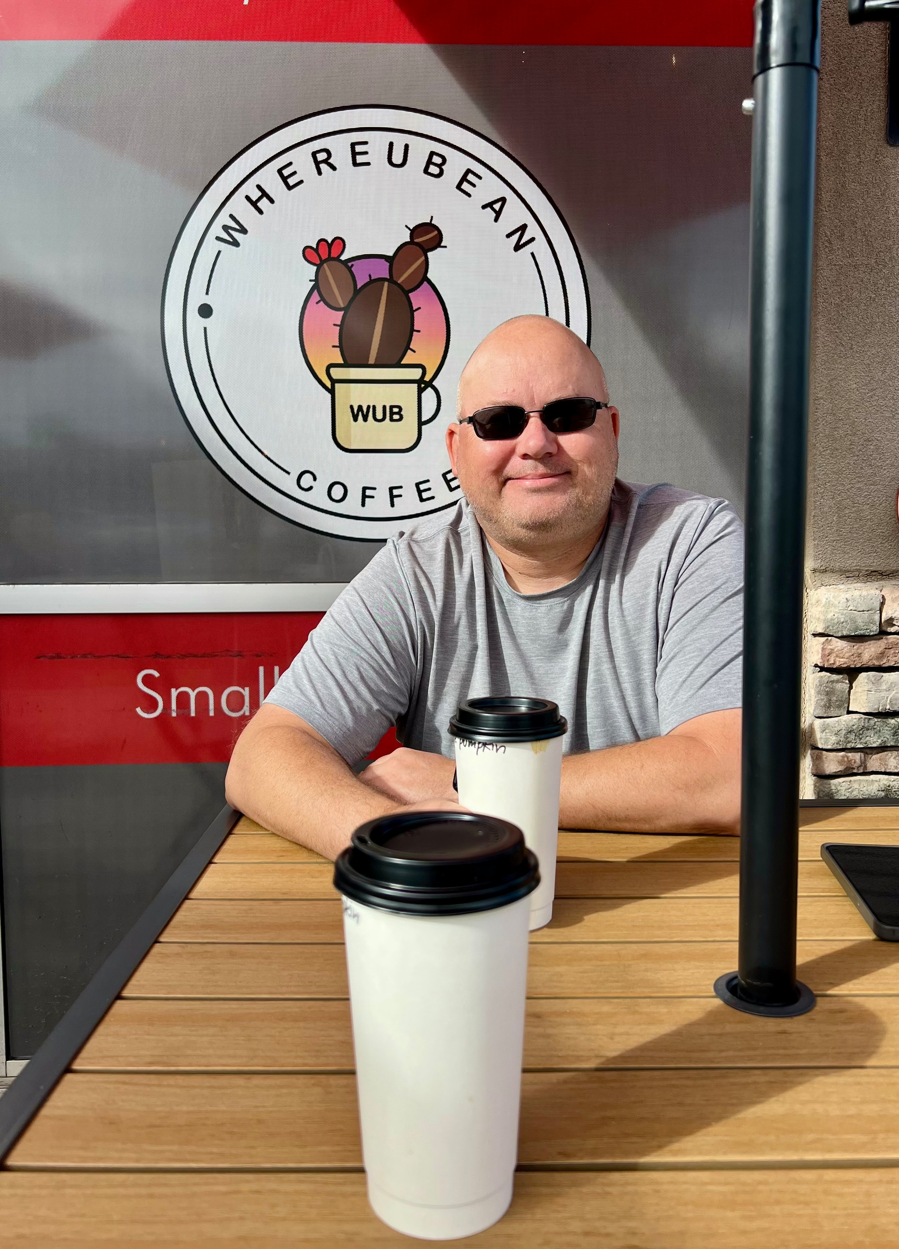 A person wearing sunglasses is sitting at an outdoor table with two takeaway coffee cups in front of a sign that reads Whereubean Coffee.