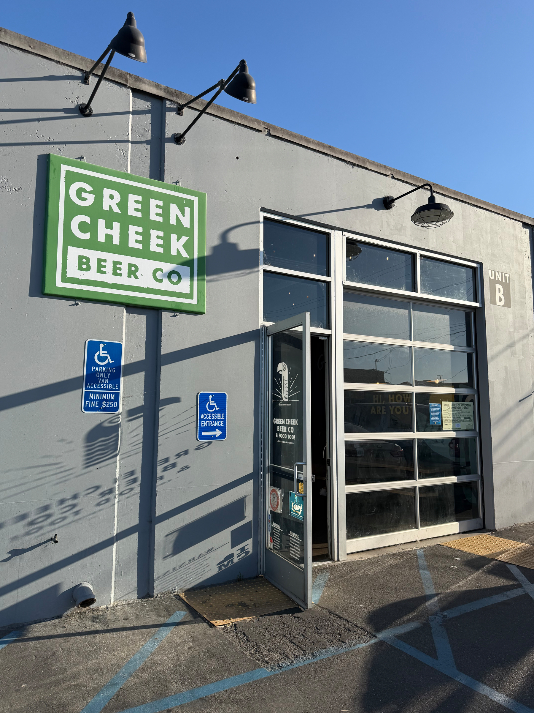 A gray building features a large glass door and a sign for Green Cheek Beer Co. under bright sunlight, with designated accessible parking spaces nearby.