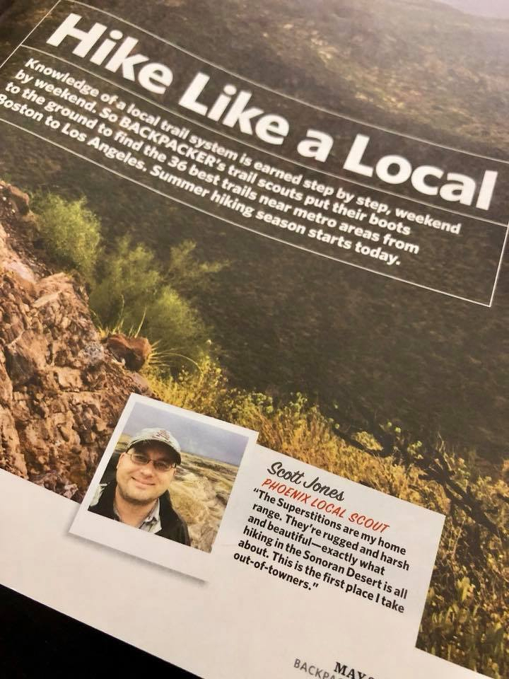 Auto-generated description: A person poses for a photo next to a magazine article titled Hike Like a Local, emphasizing hiking tips and information about different trails.